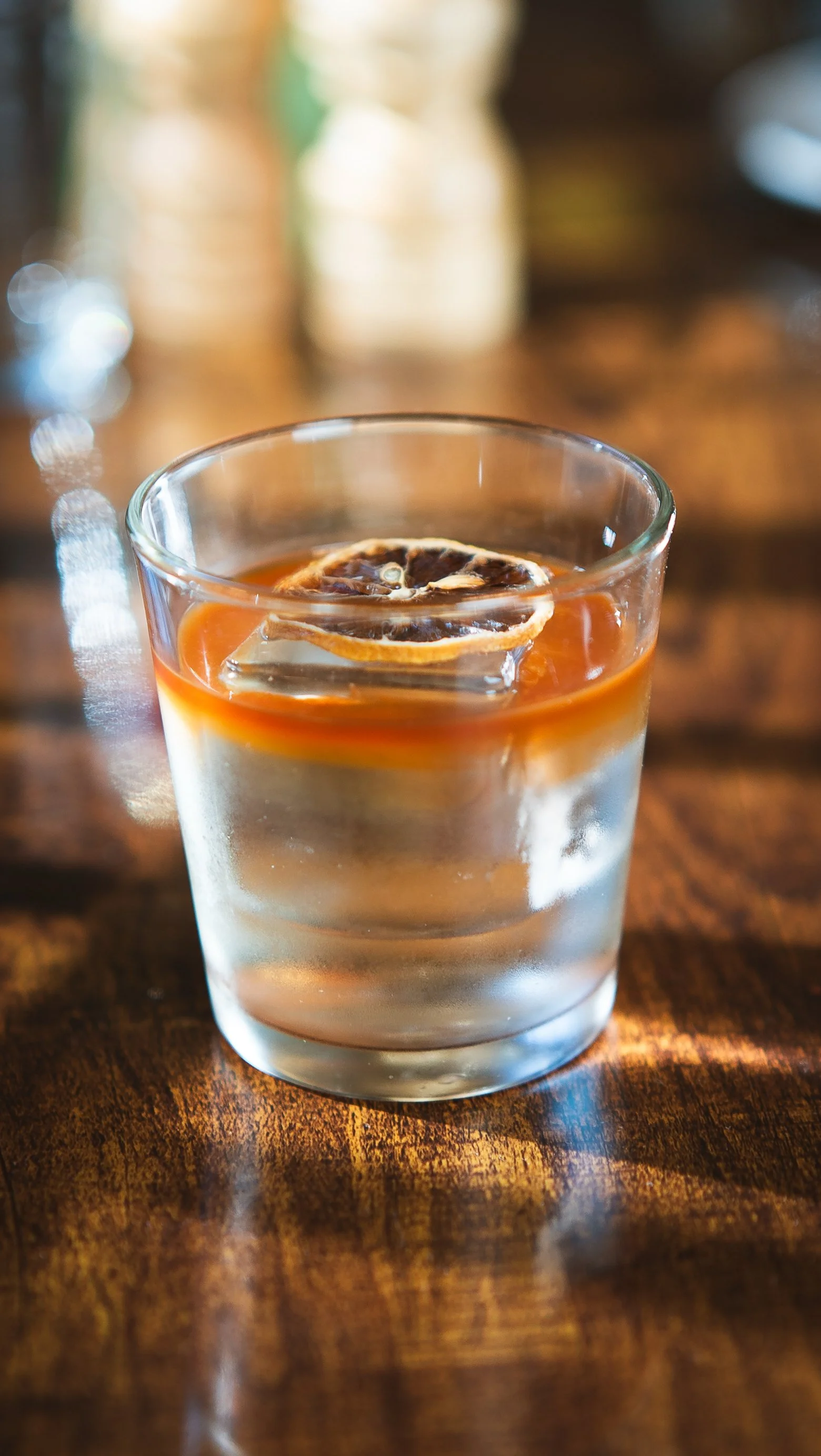 A glass of water with a slice of lemon and a slice of dried orange on top, placed on a wooden table with a blurred background.