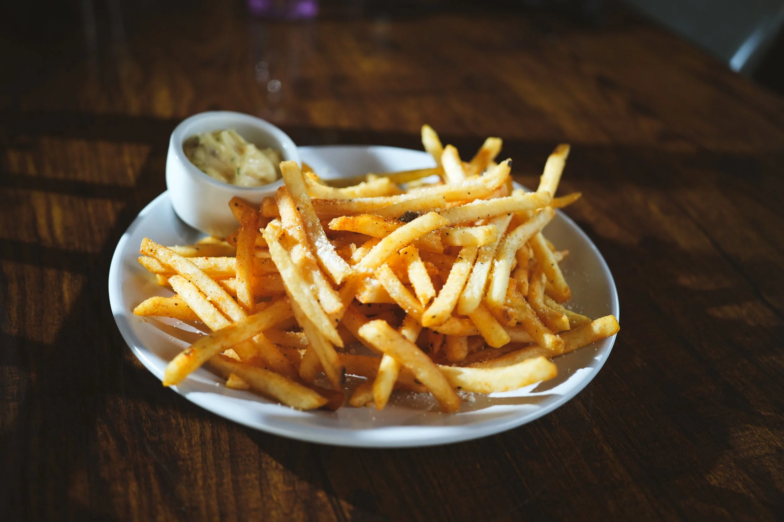 A white plate of French fries with a side of creamy dipping sauce on a dark wooden table.