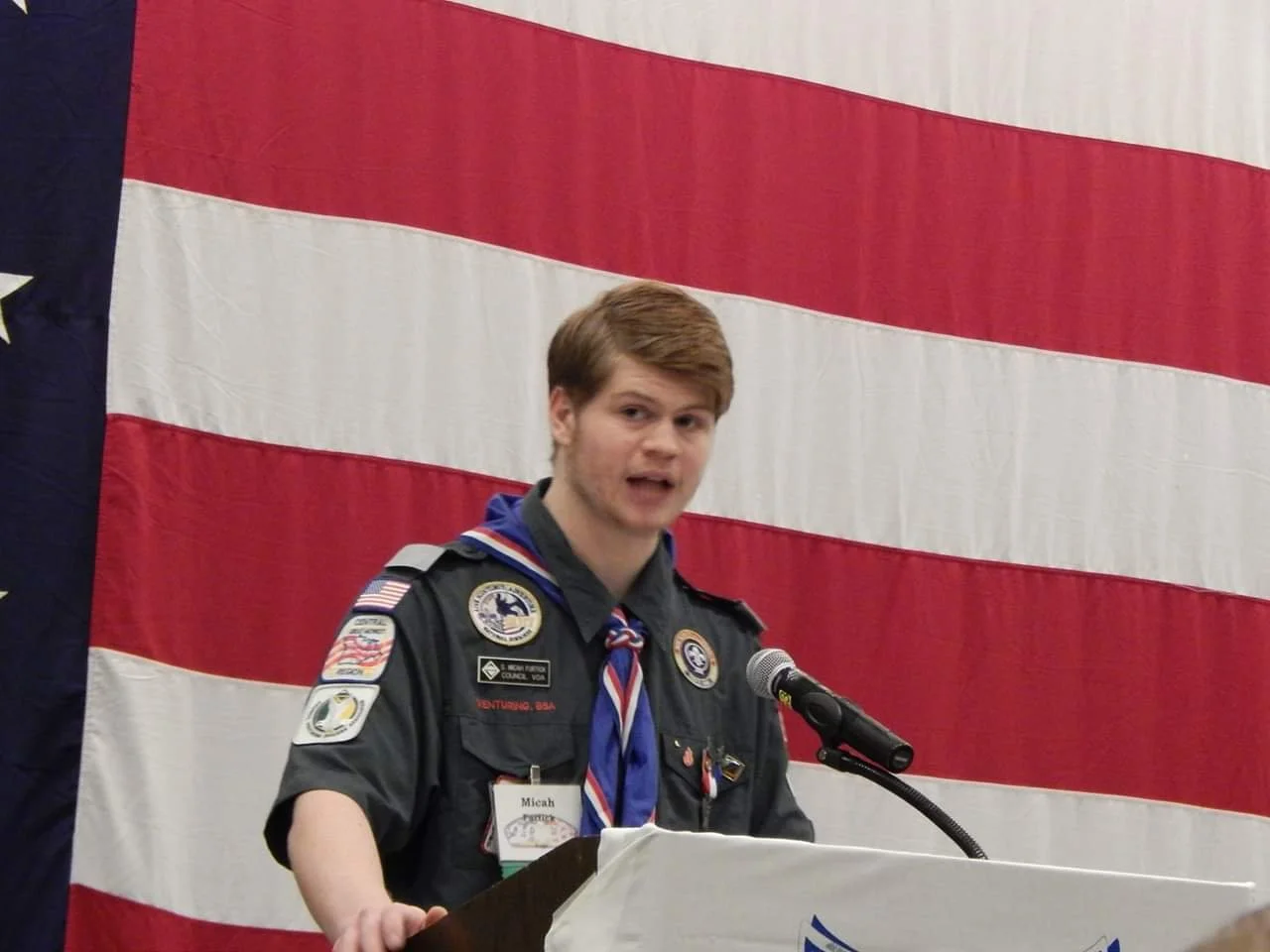 A young man in a Boy Scout uniform standing behind a podium, speaking into a microphone, with an American flag in the background.