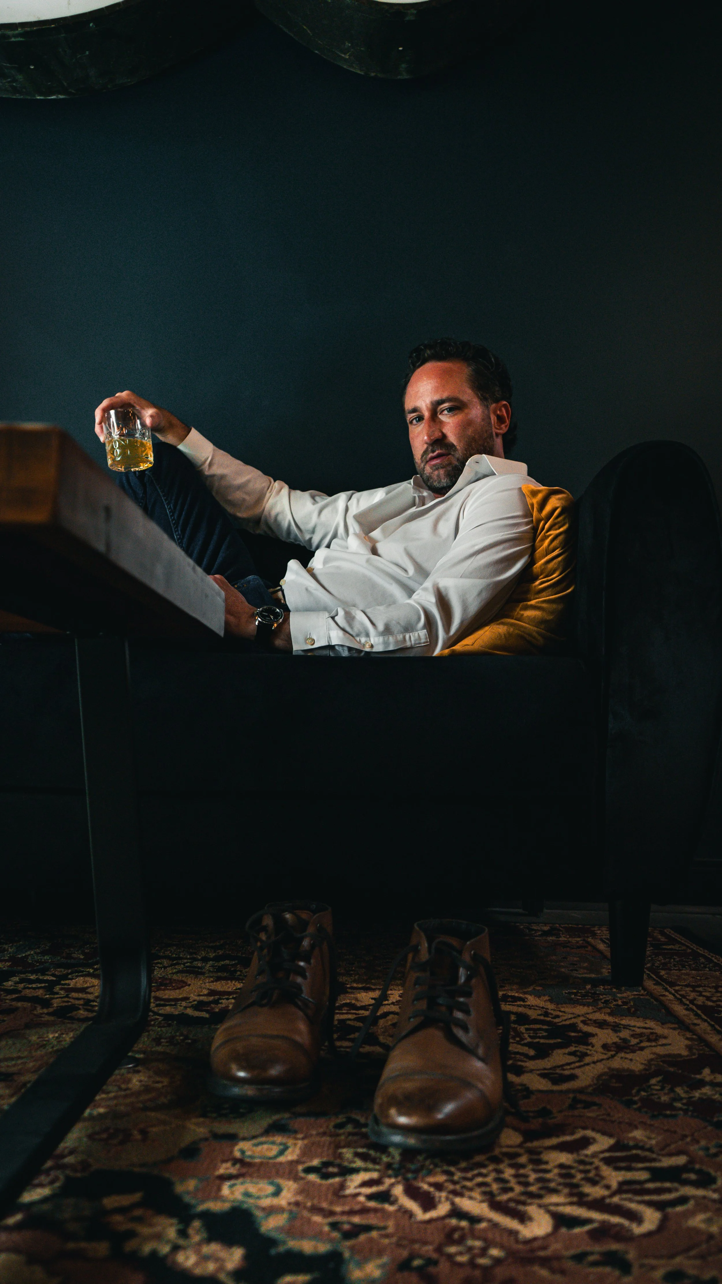 A man sits on a black couch with his feet resting on a patterned rug, holding a glass of whiskey in his right hand. He is wearing a white long-sleeve shirt, dark jeans, and brown leather shoes, with a watch on his left wrist. The background features a dark wall and a yellow pillow behind him.