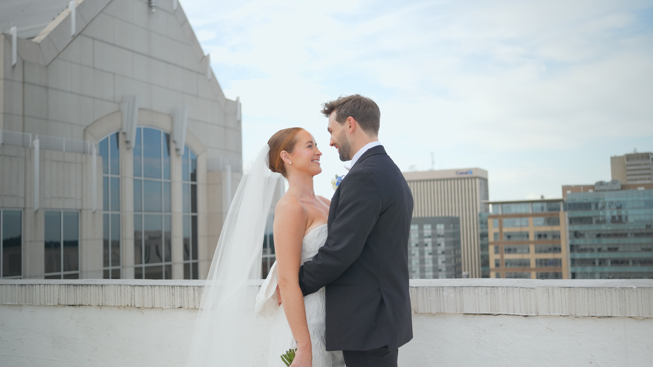 A bride and groom smile and look into each other's eyes on a rooftop with city buildings in the background, during their wedding.