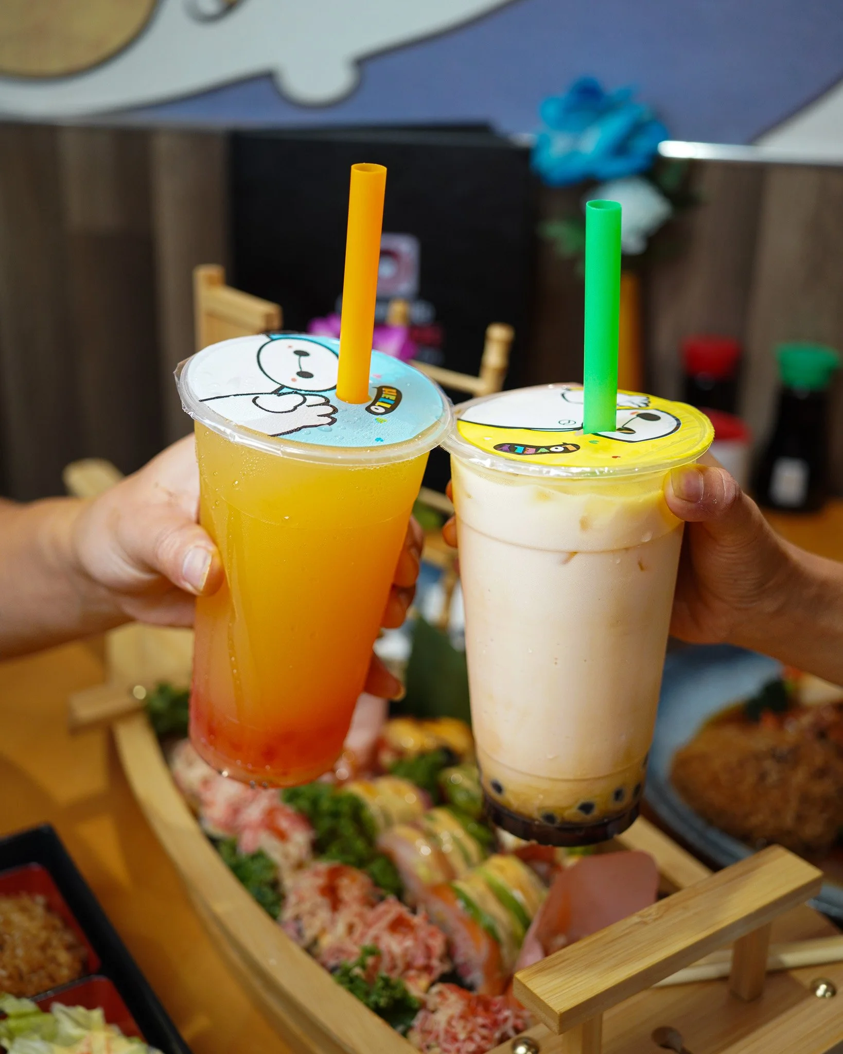 Two cups of bubble tea with colorful cartoon character lids being held above a wooden display of various sushi and other cuisines.