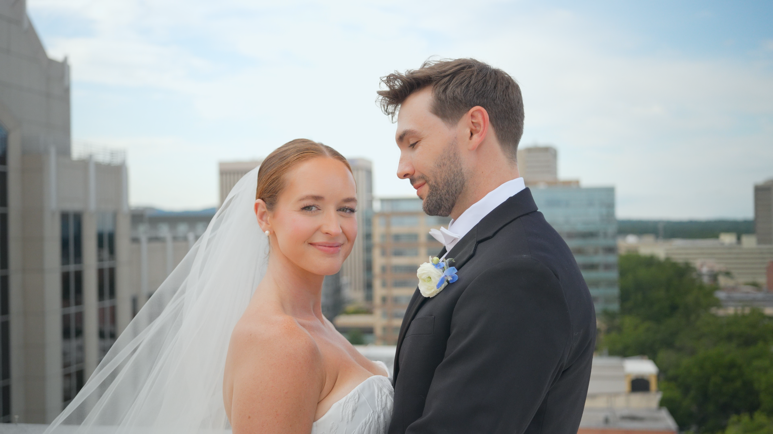A bride and groom on a rooftop in the city, standing close together and smiling, with tall buildings in the background.