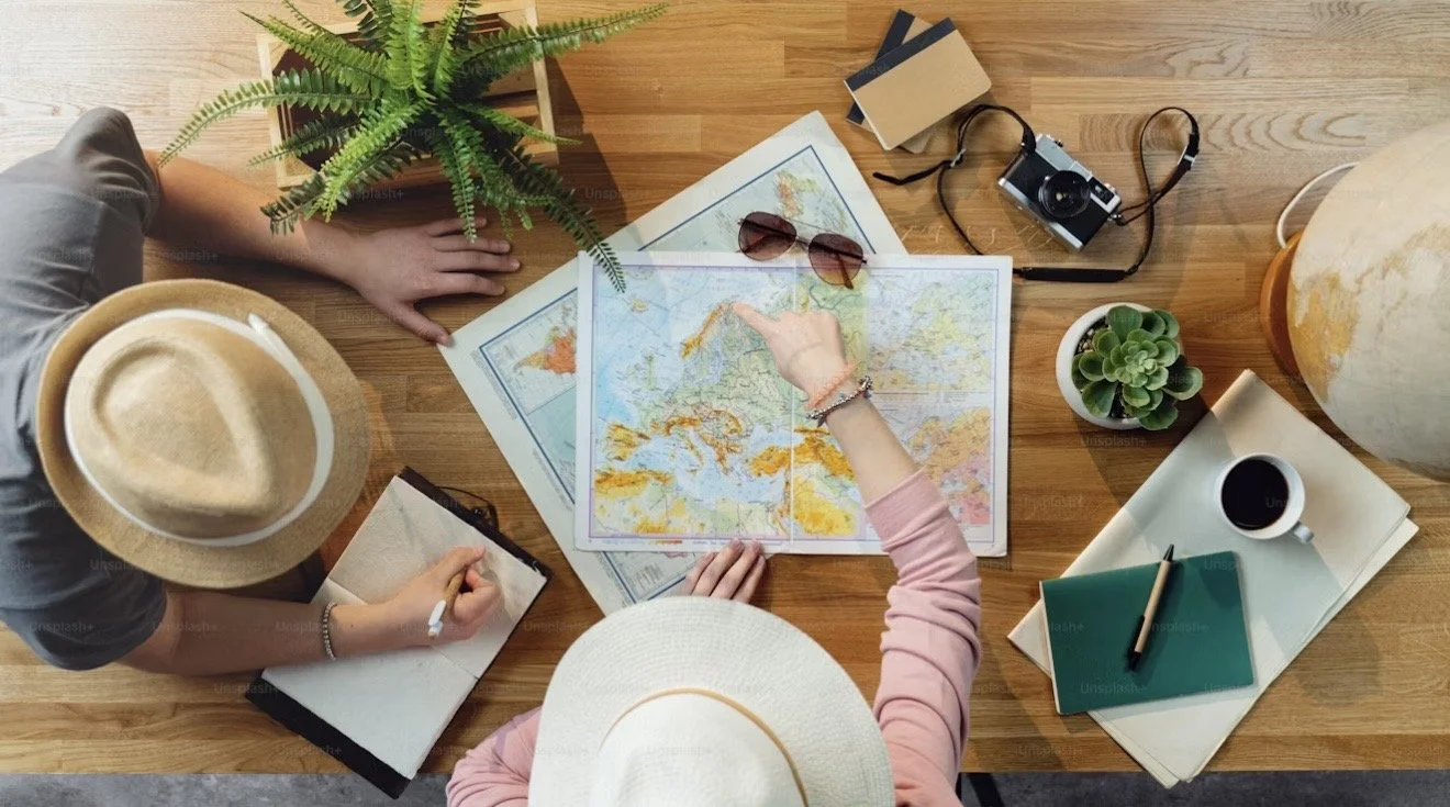 Two women with hats at a wooden table exploring a world map, surrounded by maps, glasses, a camera, potted plants, a cup of coffee, notebooks, and a globe.