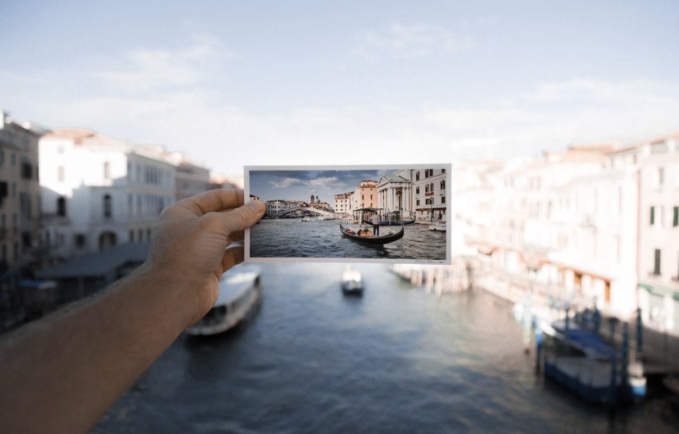Person holding a small printed photo of a canal scene with boats and historic buildings, over a blurred background of the same canal.