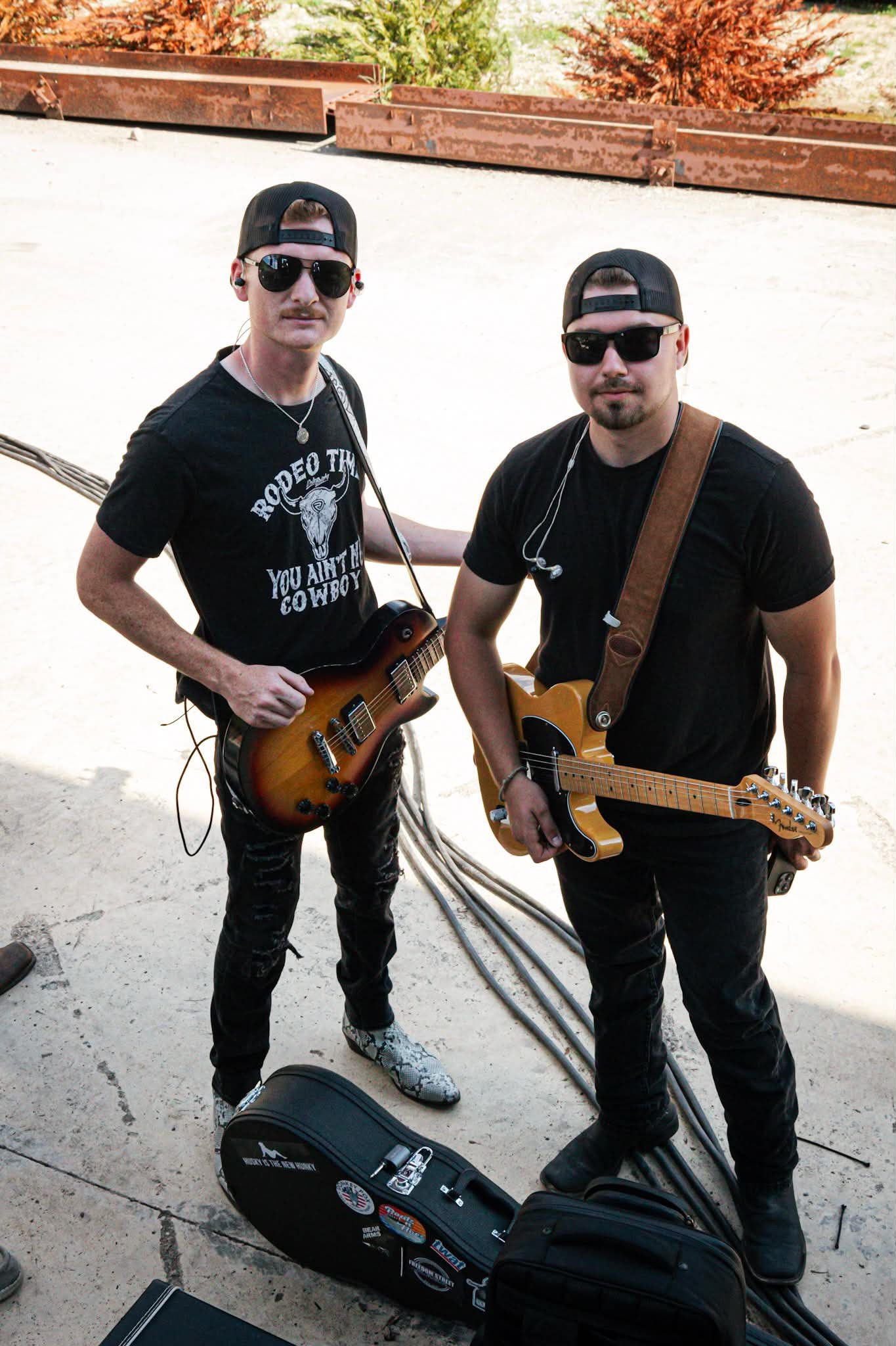 Two men wearing sunglasses, black hats, and black t-shirts with guitars around their necks standing outdoors on a sunny day. One man is holding a guitar, and there is a guitar case on the ground in front of them.