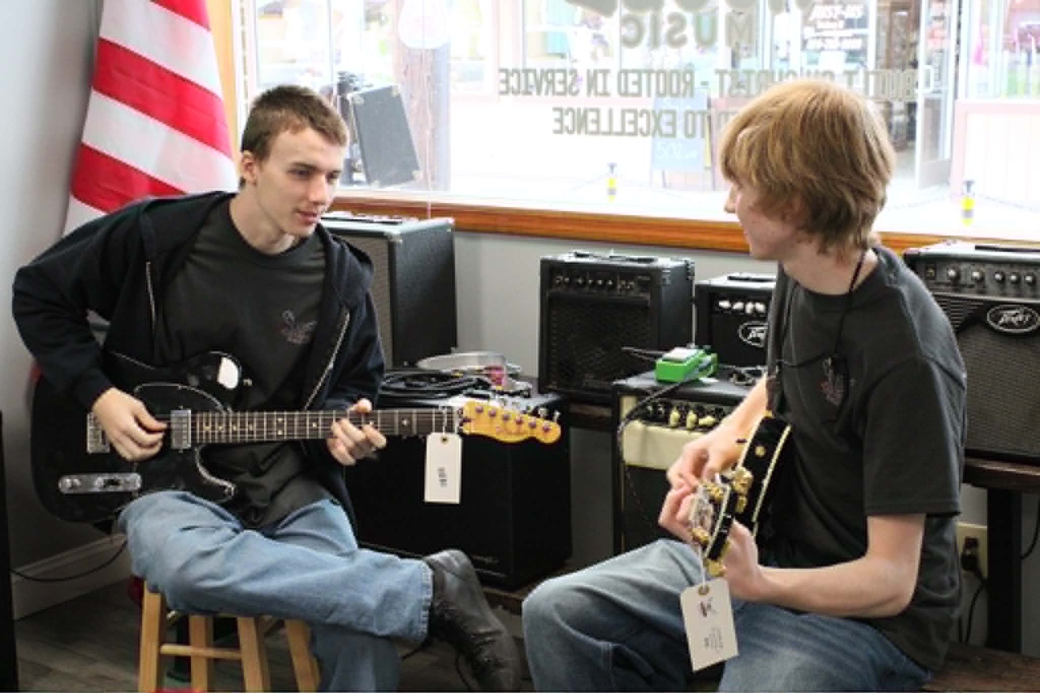 Two young men playing guitars in a music store or studio, with various musical equipment around them and a large window in the background.