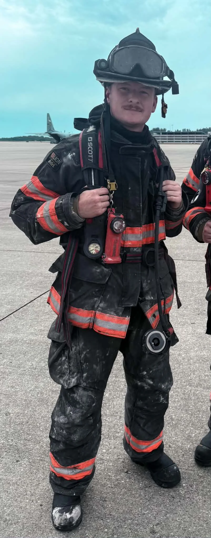 A firefighter in full gear, including a helmet, jacket, and pants with reflective stripes, standing on an airport tarmac with a plane in the background.