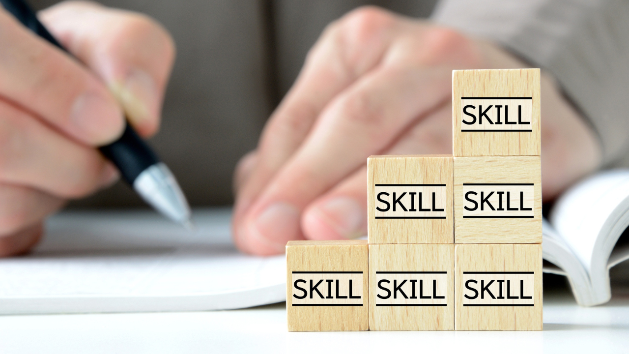 Wooden blocks stacked to form a pyramid with the word 'Skill' on each block, with a person's hand writing nearby.