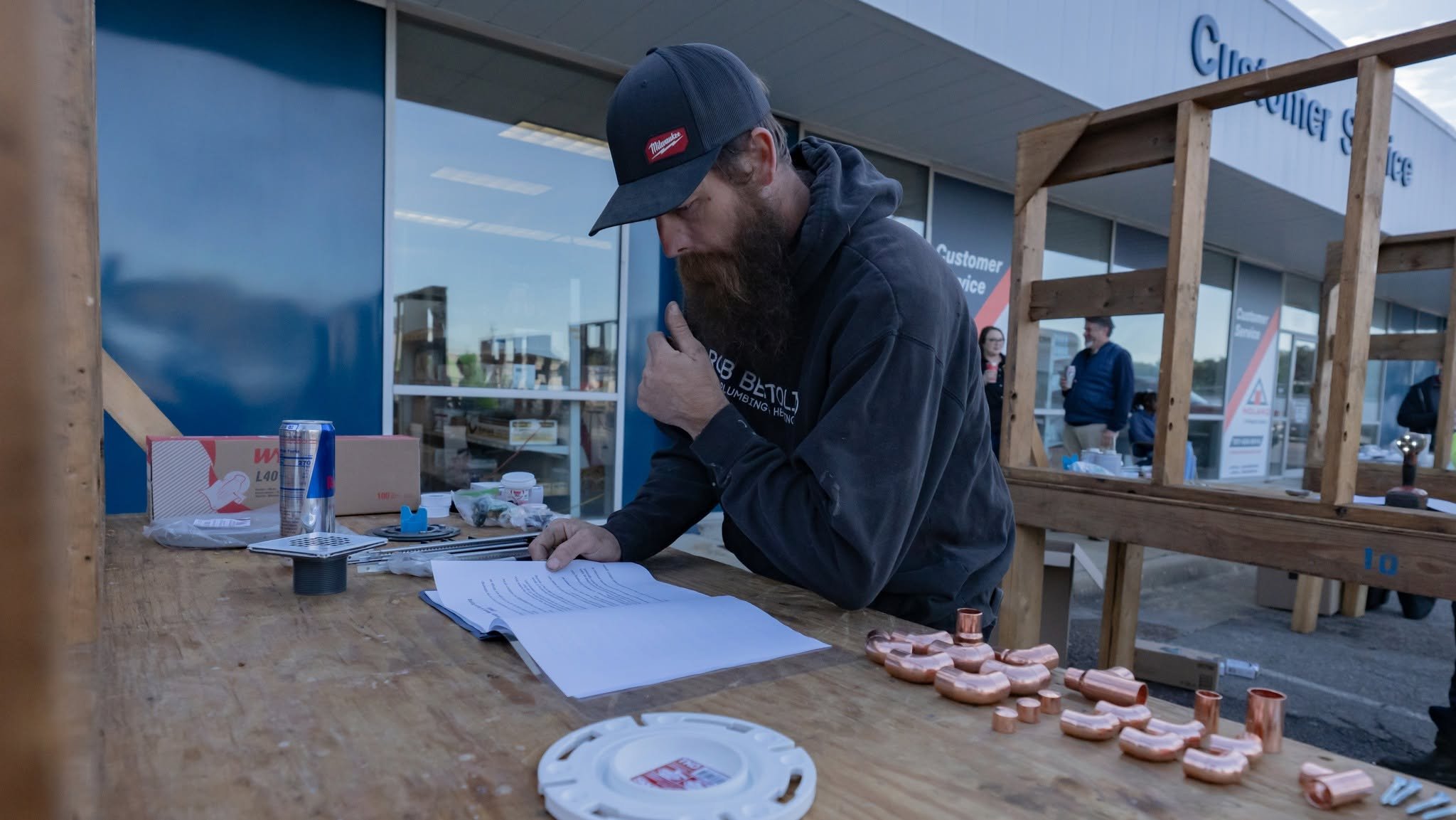 A man with a beard wearing a cap and black hoodie is working at an outdoor table with various plumbing parts, paperwork, and a soda can. There are other people in the background standing in front of a building with glass windows.