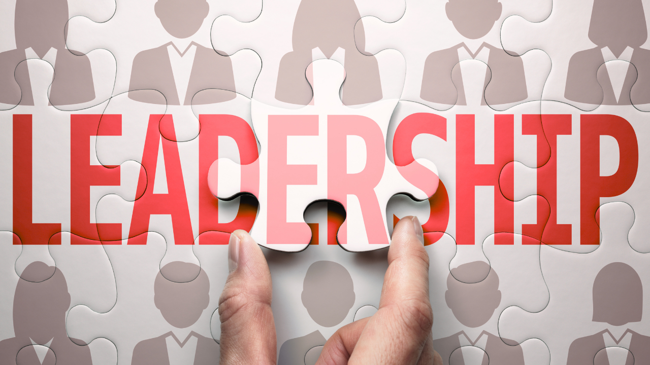 Puzzle pieces with the word 'LEADERSHIP' in bold red letters, a hand placing a piece in the puzzle, background shows silhouettes of diverse people.