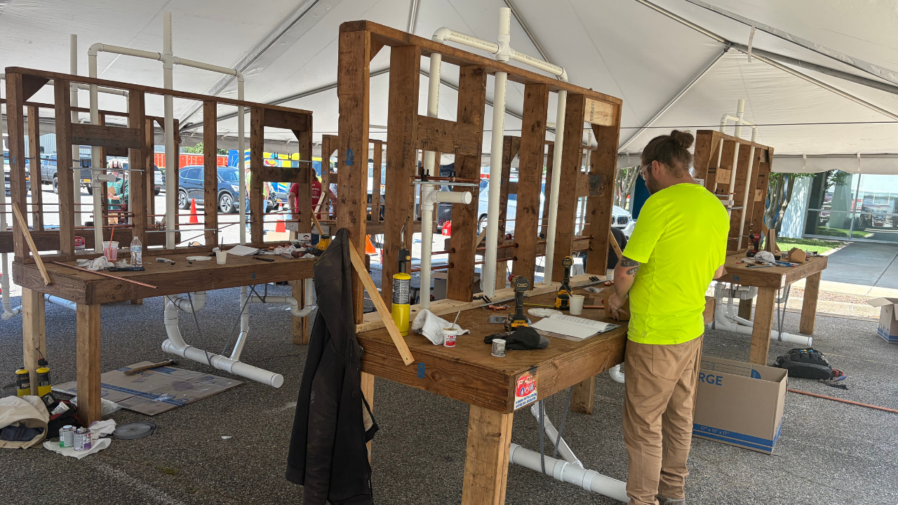 Person working on a construction project inside a large white tent with makeshift workbenches made from wooden pallets and PVC pipes for an apprenticeship competition.