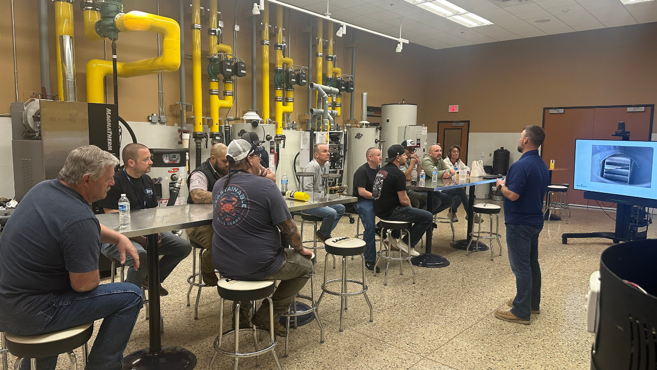 Man giving a presentation to a group of people sitting at a long table in a room with industrial equipment and yellow pipes in the background.