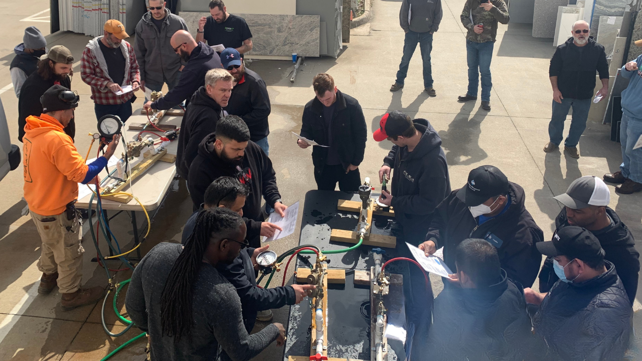 A group of people gathered outdoors around work tables with equipment, some holding papers, others working on assembled backflow preventers.