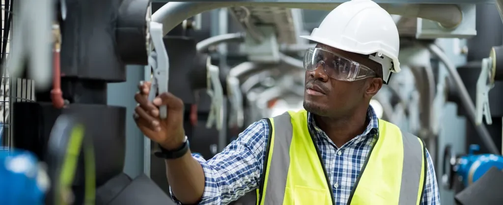 A man wearing a white safety helmet, clear safety glasses, a high-visibility vest, and a checkered shirt inspects machinery in an industrial setting.