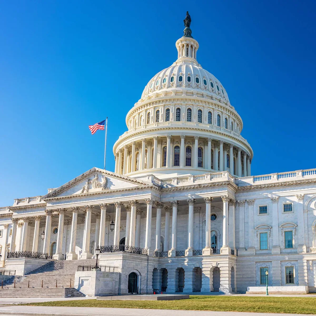 The United States Capitol building with a blue sky in the background, featuring a prominent dome and an American flag flying nearby.