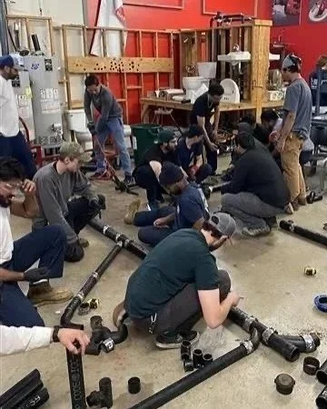 A group of people assembling cast iron black pipes and fittings on the floor in a workshop or classroom setting for Charlotte Pipe and Foundry, learning how to weld.