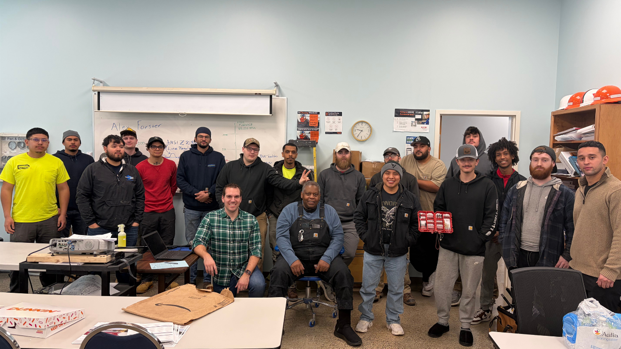 Group of around 16 people gathered in a classroom, some standing and some sitting, after a plumbing training at a trade school.
