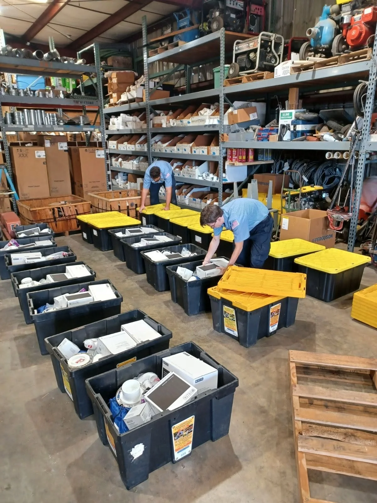 Two workers in a warehouse organizing black storage bins filled with various supplies, with shelves stocked with boxes, pipes, and equipment in the background.