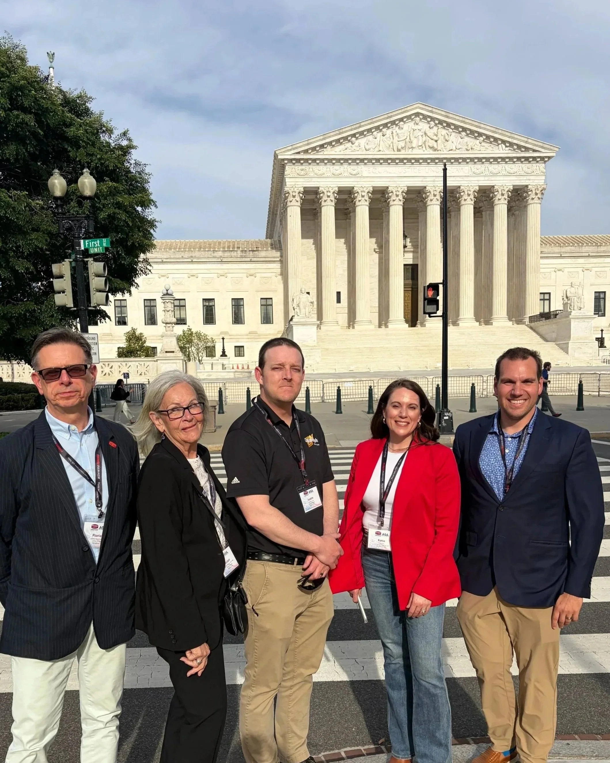 Group of six people standing in front of the U.S. Supreme Court building in Washington, D.C., with the steps and columns visible behind them. The group is smiling and wearing conference badges.