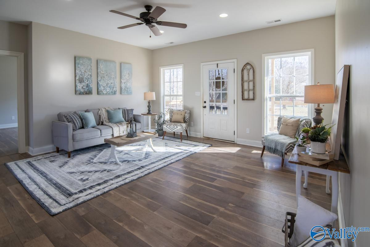 Bright living room with a beige sofa, decorative pillows, and a framed abstract art piece on the wall. Includes a patterned armchair, side table with a lamp, a large window, and a door leading outside. Hardwood floors and a ceiling fan complete the s