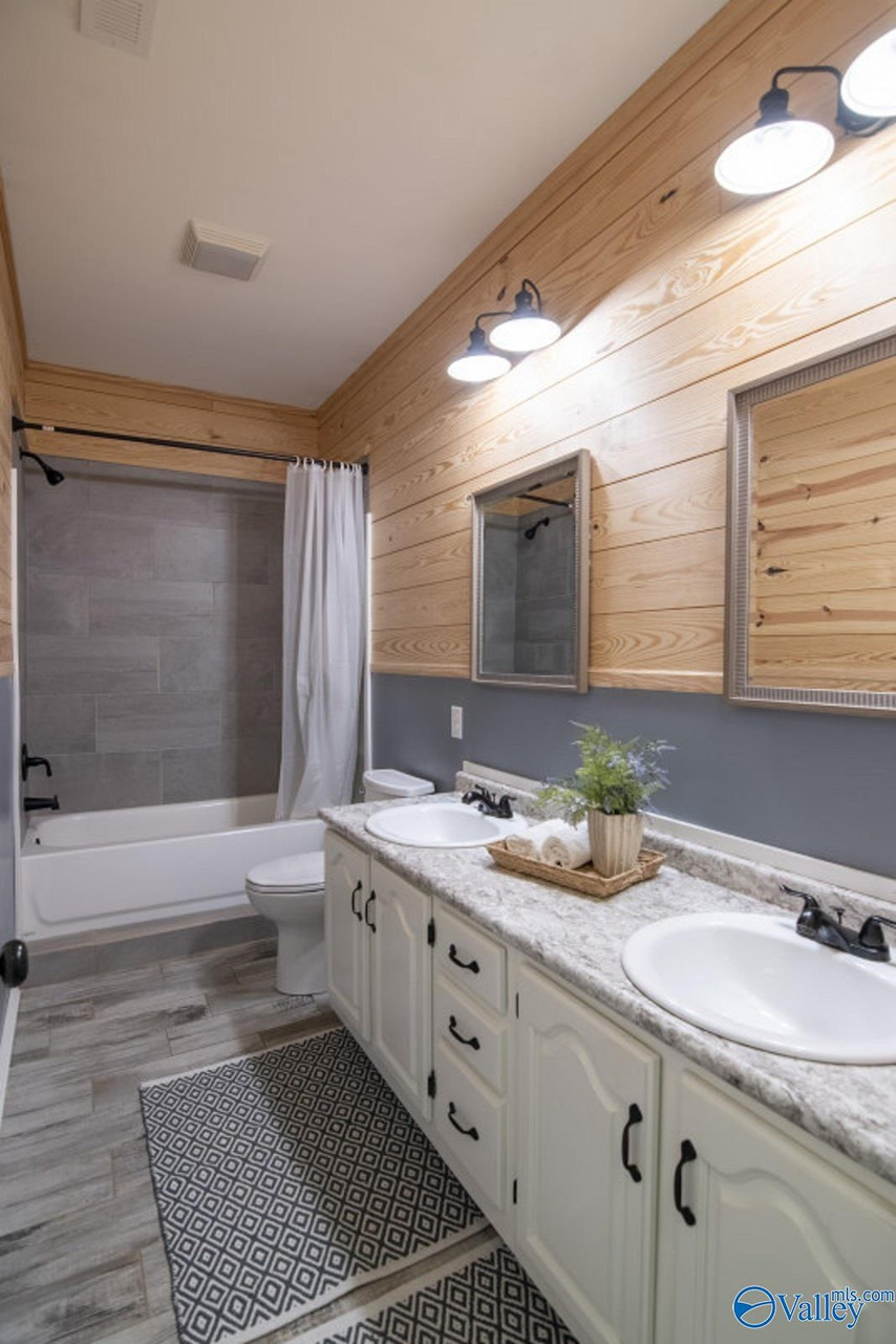 A bathroom with a double vanity, two oval sinks, black fixtures, a granite countertop, and two mirrors. There are wooden accents on the wall, two wall-mounted light fixtures, a bathtub with a gray tile surround, a white shower curtain, and a patterne