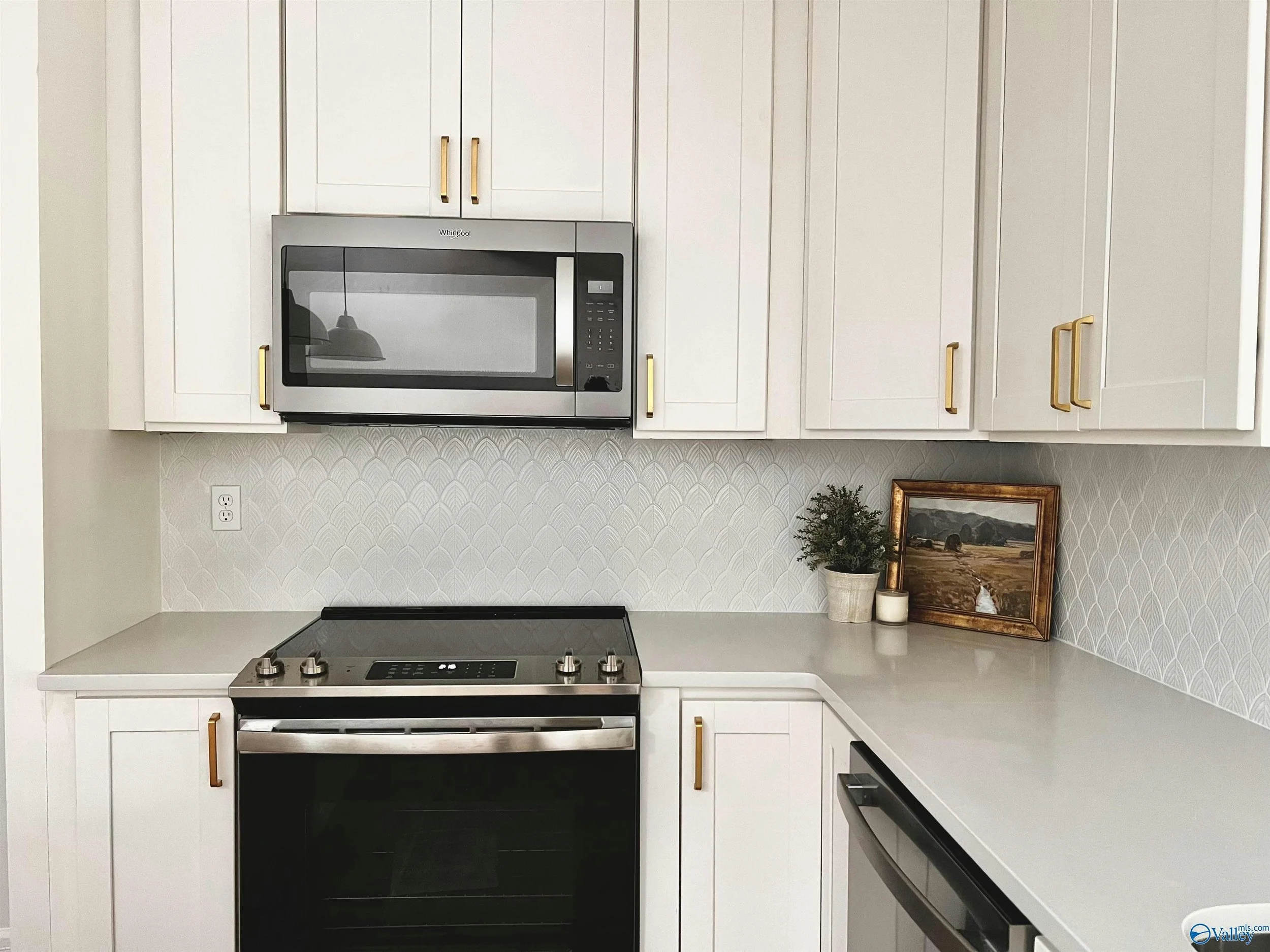 White kitchen cabinets with gold handles, a stainless steel microwave oven, a stove, a framed landscape photo, a small plant, and a candle on the counter.