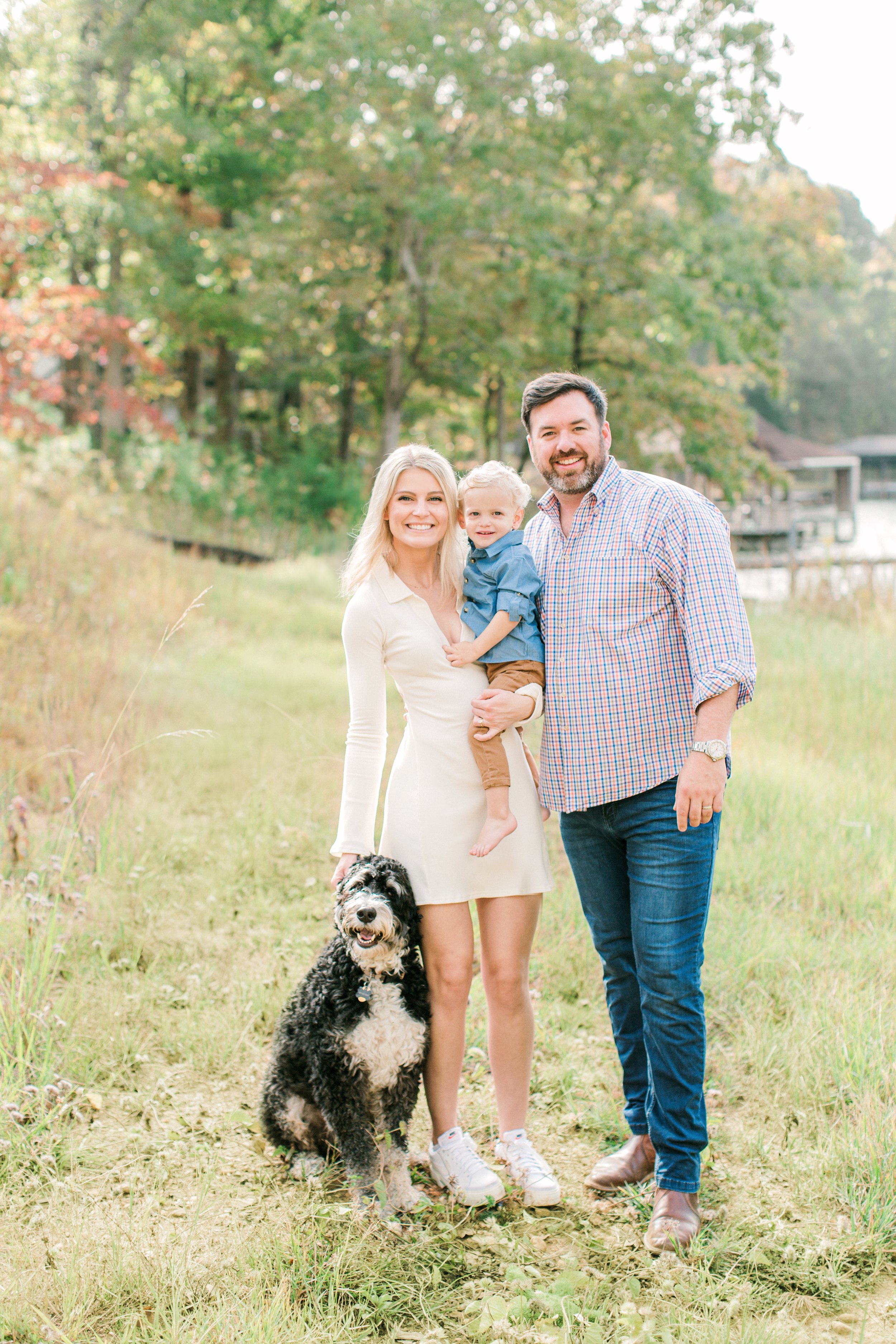 A smiling family of three and their dog outdoors in a grassy park with trees in the background during daytime.
