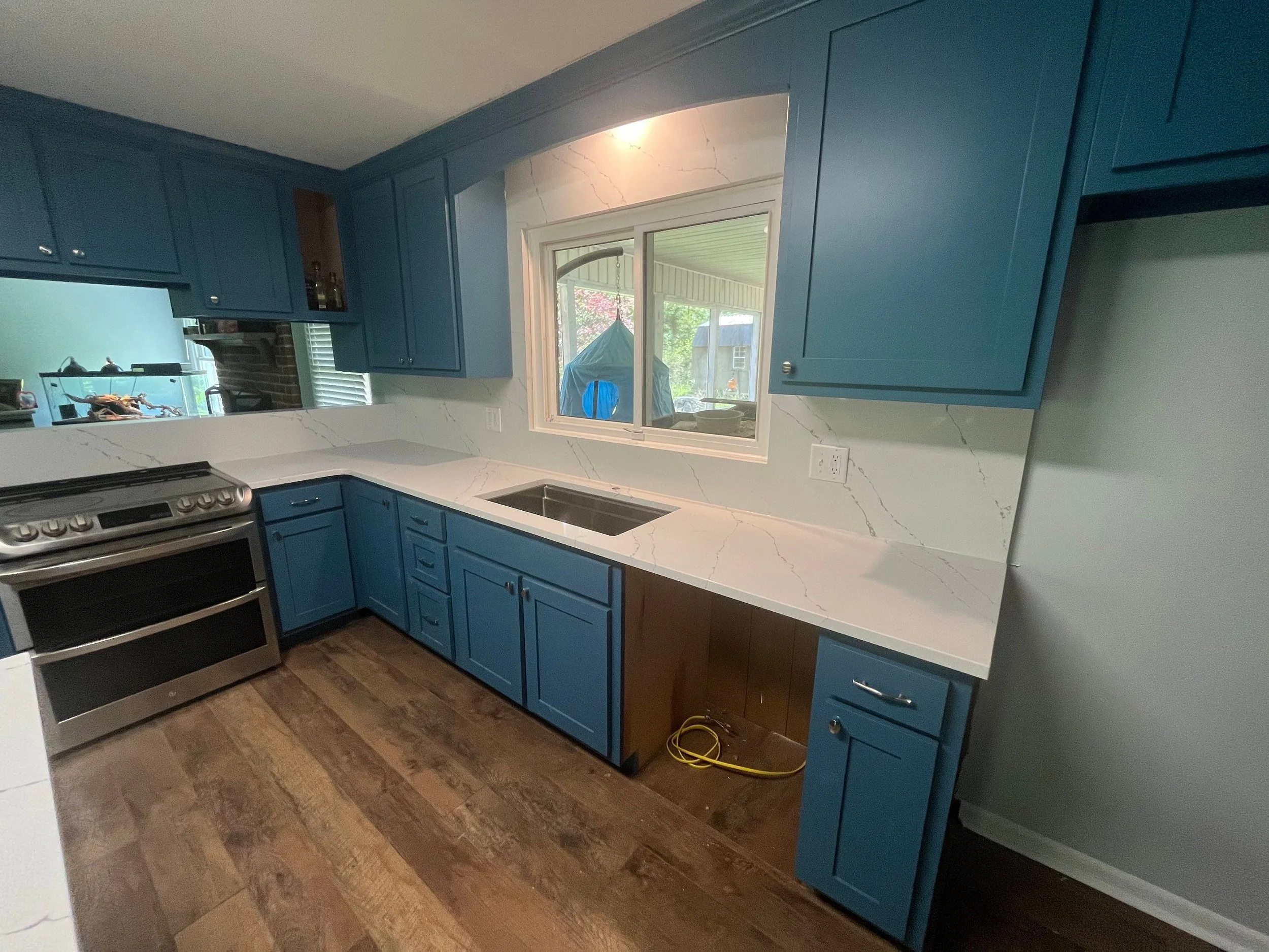 The image shows a kitchen with blue cabinets, white marble countertops, a stainless steel stove, a window above the sink, and a view of a screened porch with outdoor furniture outside.