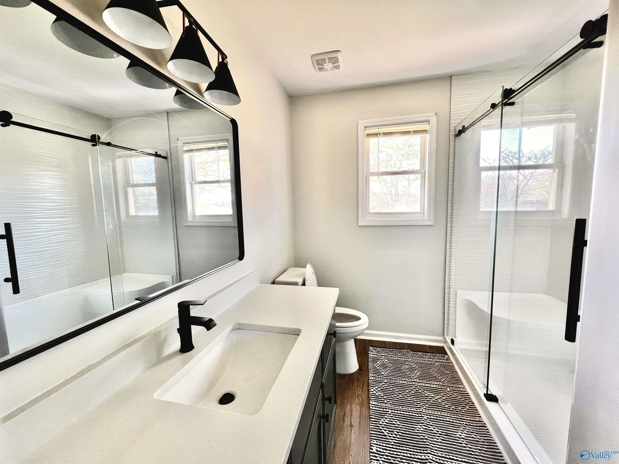 A bathroom with a white vanity, black faucet, and large mirror. There are two windows with blinds and a glass shower with a sliding door. The floor has a black and white striped rug.