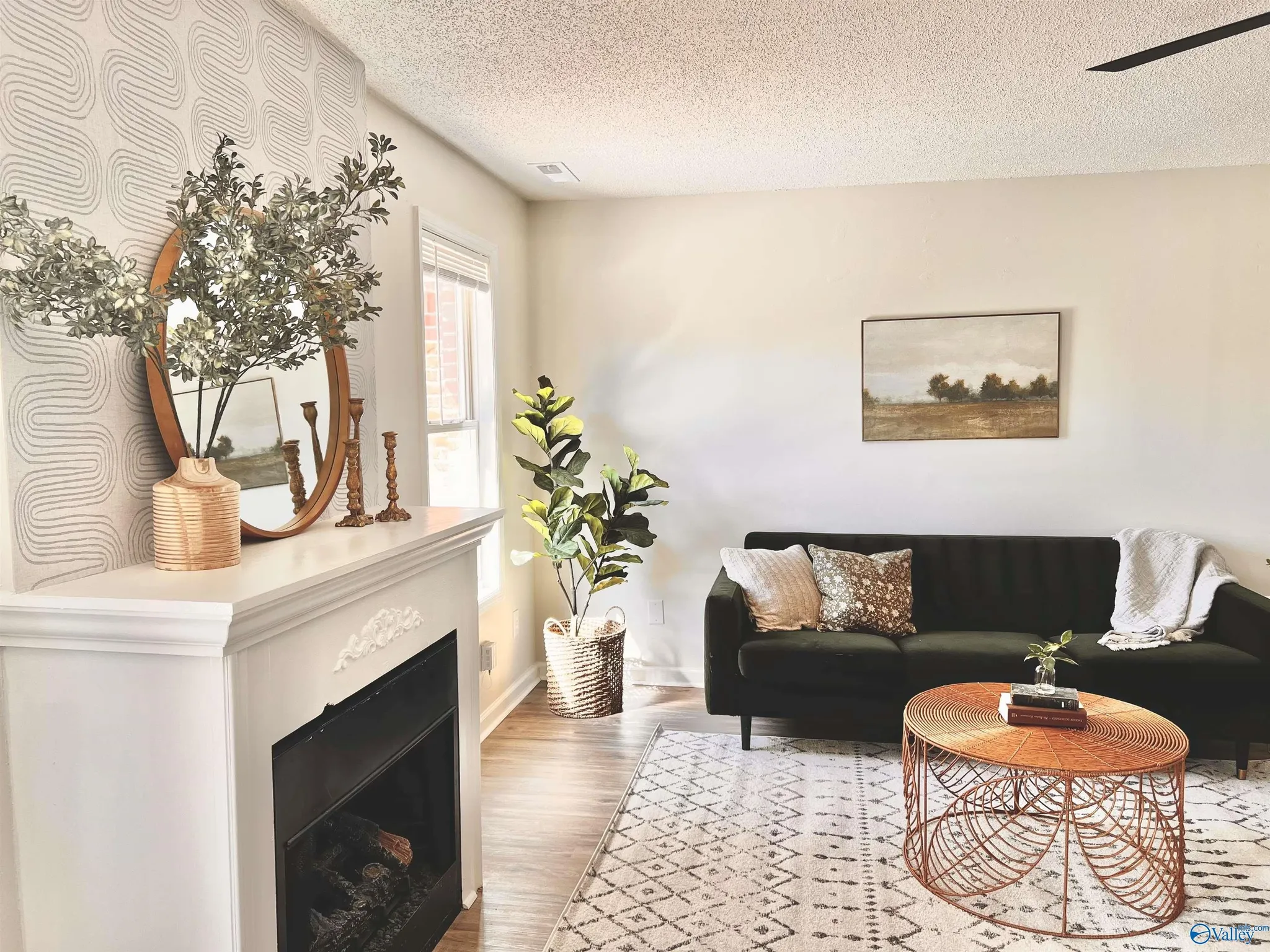 Living room with a black sofa, a round orange wire table, a rug with black diamond pattern, a white mantel with decorative items, a window with blinds, a framed landscape painting, and indoor plants.