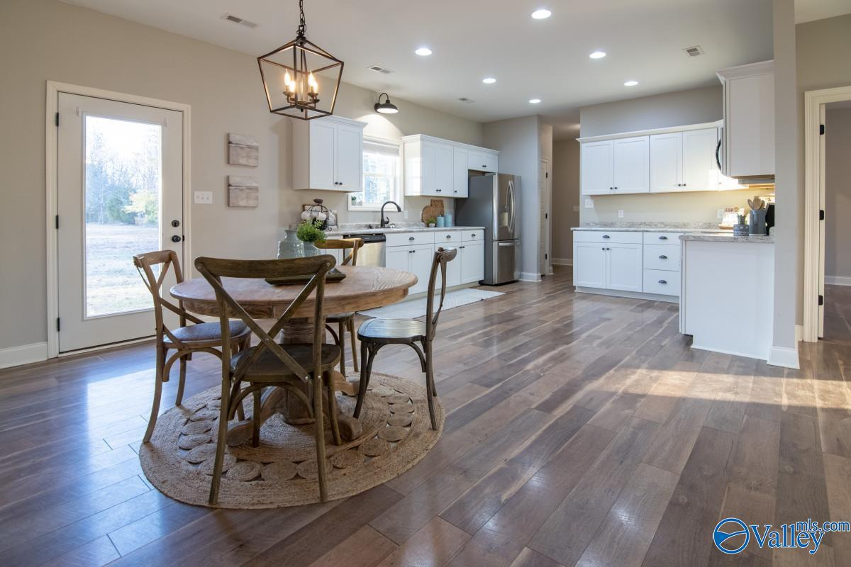 Bright, modern kitchen with white cabinets, stainless steel appliances, and wooden flooring. A round dining table with four chairs is situated in the foreground, on a woven rug. There is a door with a window on the left side.