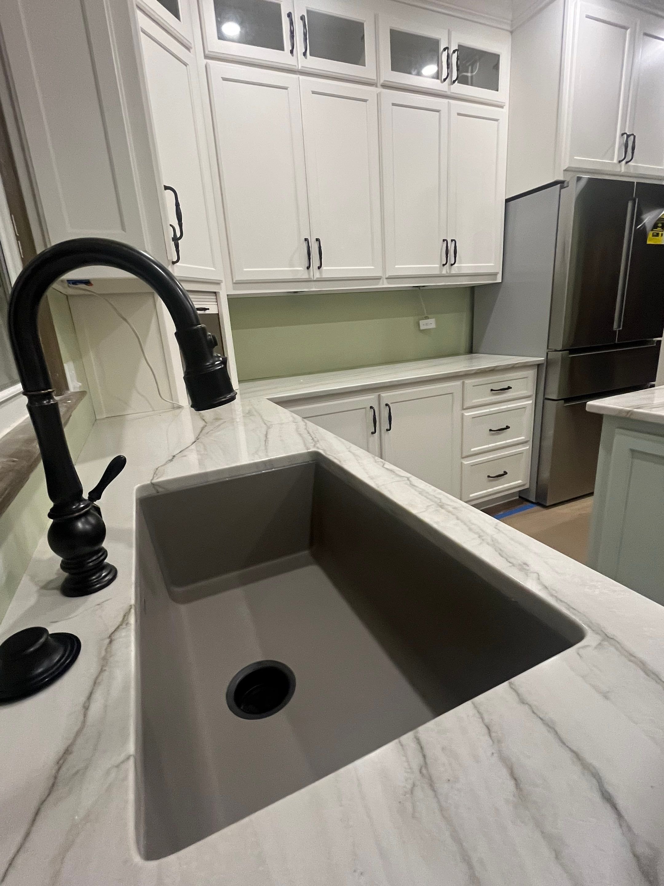 Kitchen sink with a black faucet and white marble countertop, white cabinets with black handles, stainless steel refrigerator, and a green accent wall.