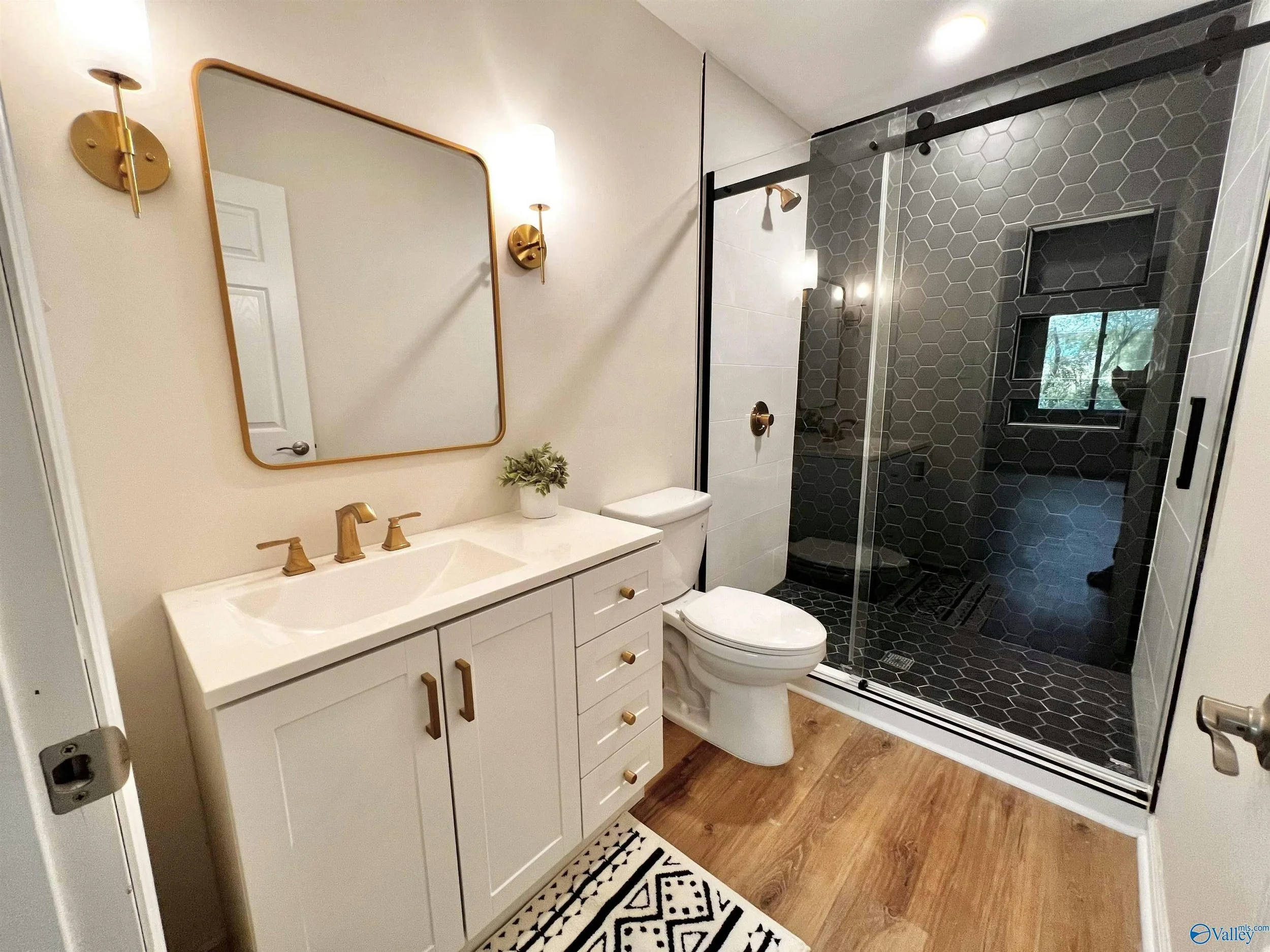 A modern bathroom with a white vanity with gold handles, a large mirror, and two gold wall sconces. Next to the vanity is a toilet, and in the background is a shower with black hexagon tiles, a glass door, and a built-in niche. The bathroom has woode