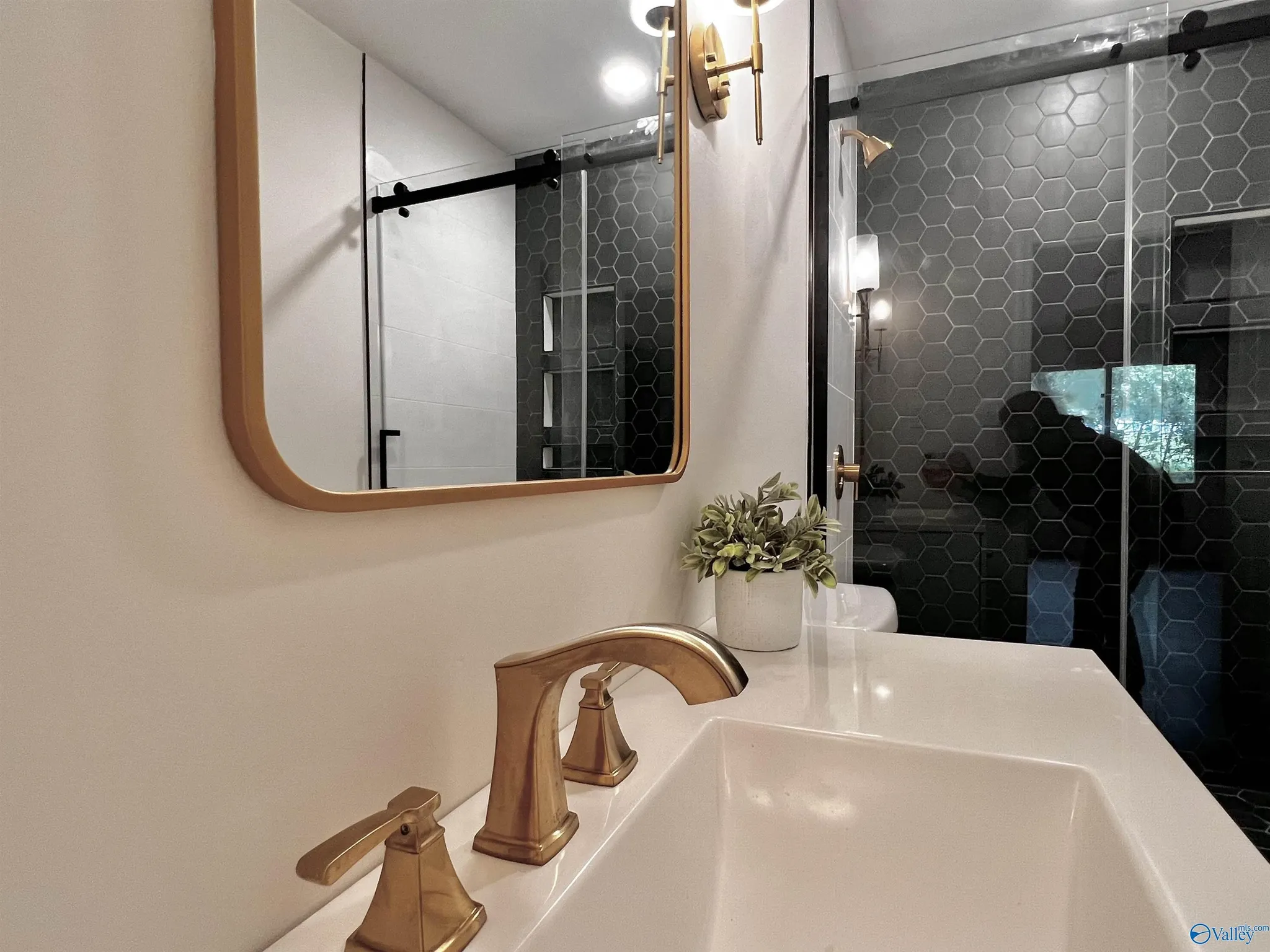 A bathroom sink with a gold-colored faucet and soap dispenser. A mirror above the sink reflects a shower area with a hexagon tile wall and black sliding door. There is a small potted plant near the sink.