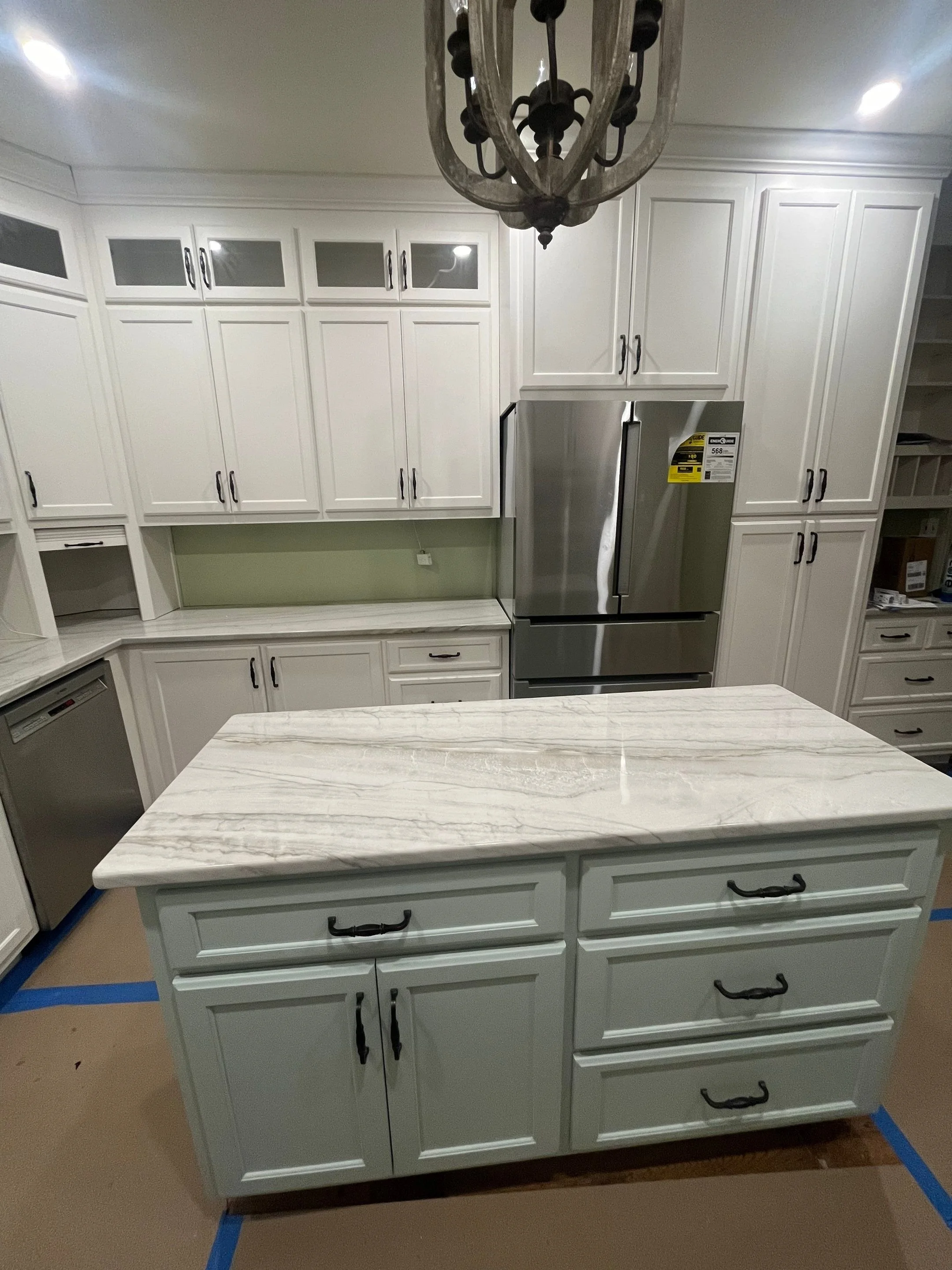 Kitchen with white cabinets, a marble countertop, and a stainless steel refrigerator. There is a chandelier hanging from the ceiling. The floor is covered with brown paper and painter's tape.