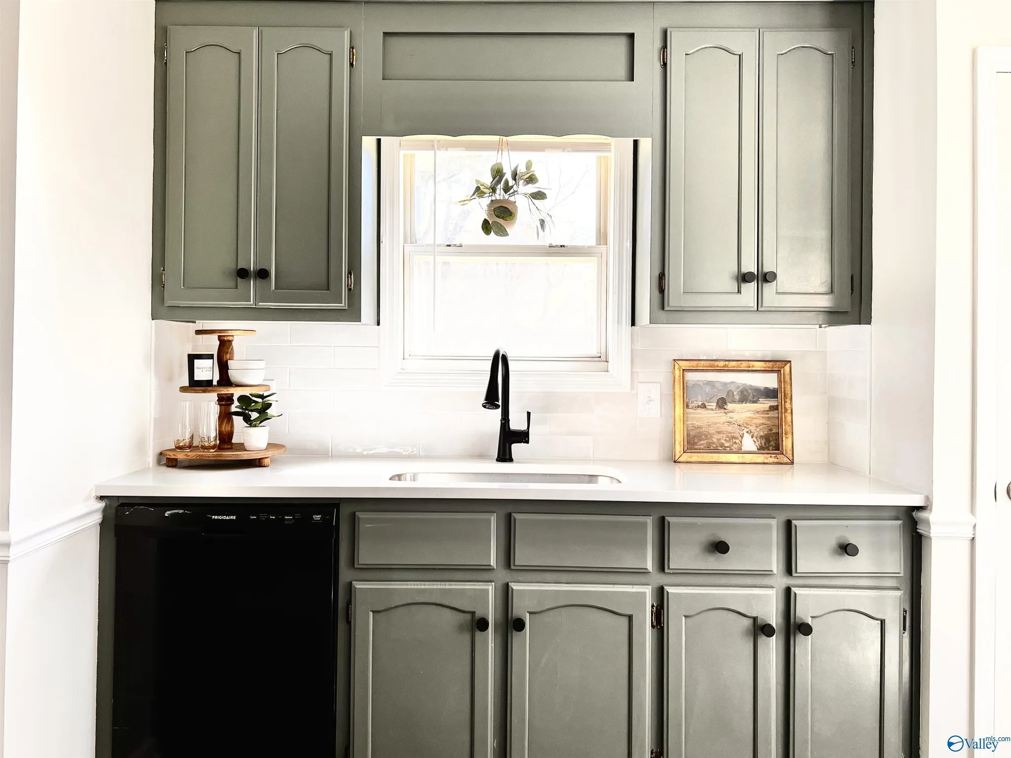 Kitchen with gray cabinets, a window with a hanging plant, a small shelf with a potted plant, and framed artwork. Black sink faucet, dishwasher, and white backsplash.