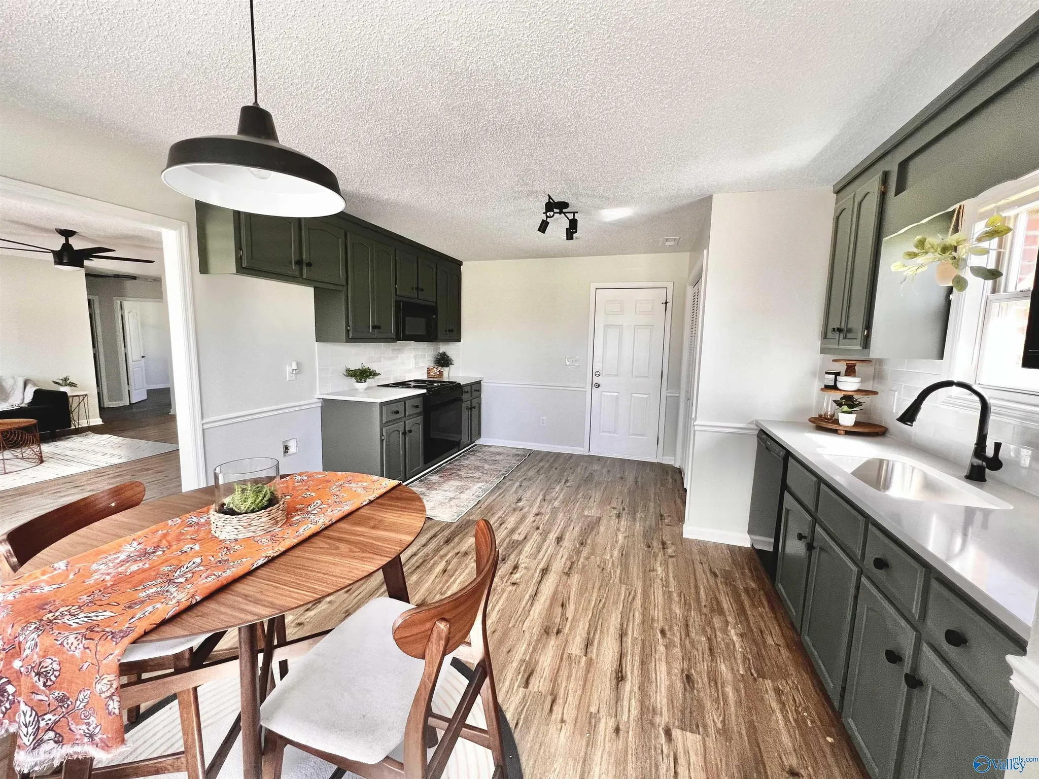 Interior of a kitchen with gray cabinets, white countertops, and a stainless steel sink. There is a dining table with a colorful orange and white tablecloth and wooden chairs. A window with a small plant is above the sink. The kitchen has wooden floo