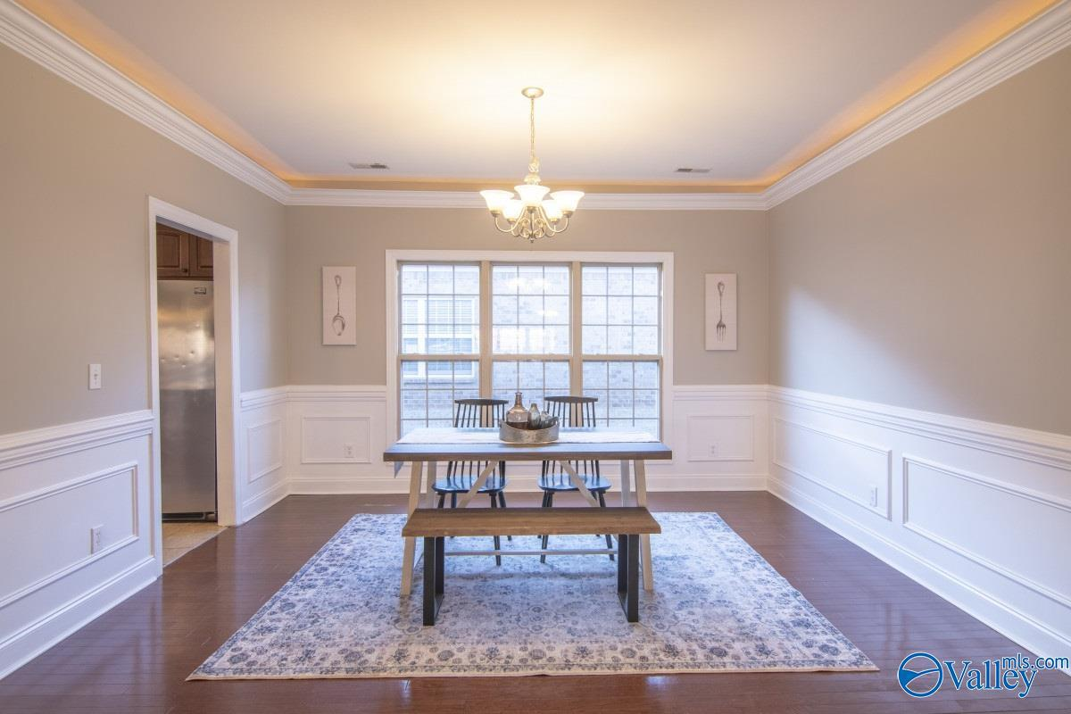 Dining room with a wooden table, four chairs, a chandelier, and large window with blinds. Decorative artwork hangs on the walls, and a patterned rug is underneath the table.