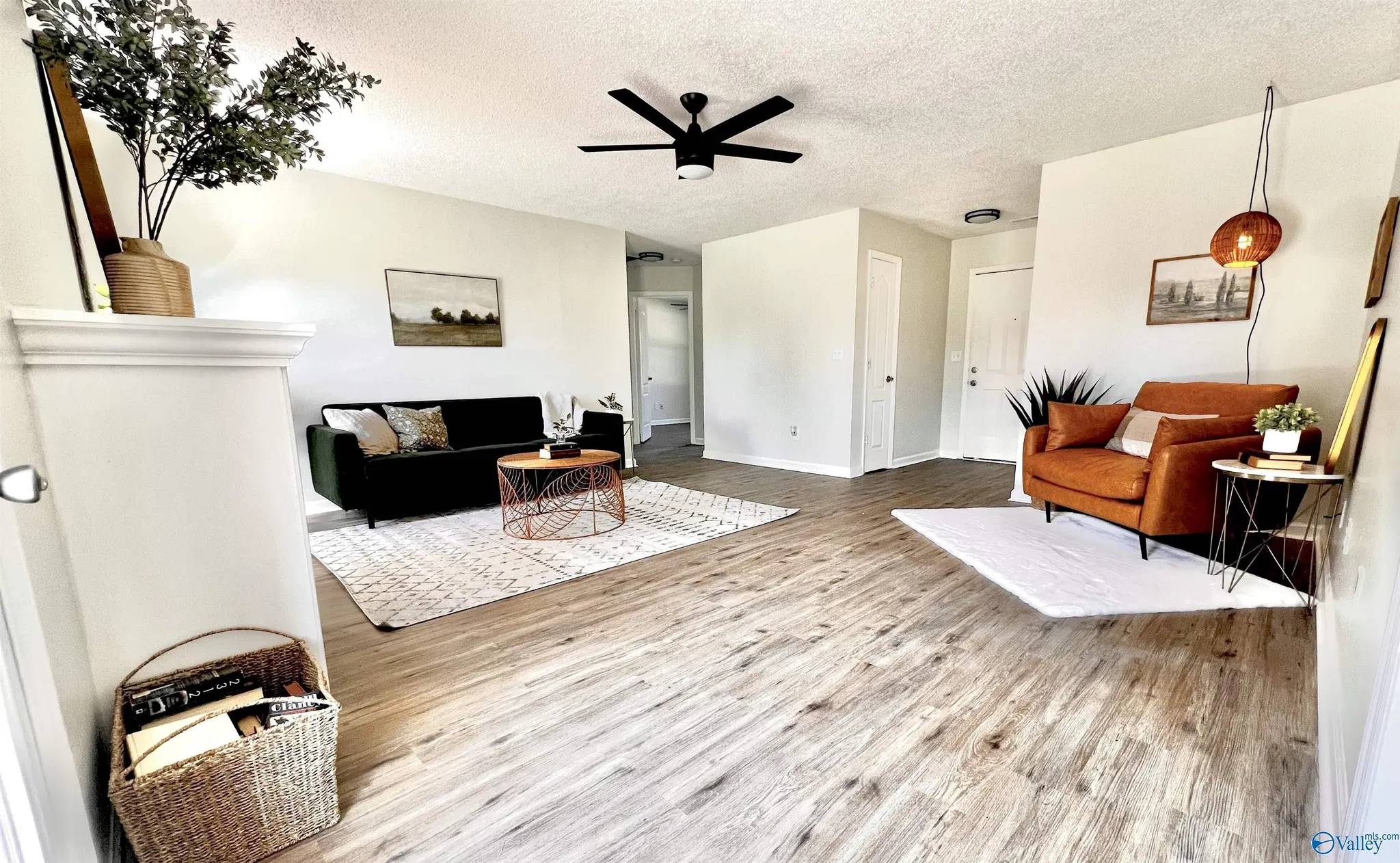 A living room with white walls, wooden flooring, a black couch with decorative pillows, a wooden coffee table, a rust-colored armchair, a small side table with plants, framed artwork on the walls, a ceiling fan, and a hanging lamp.