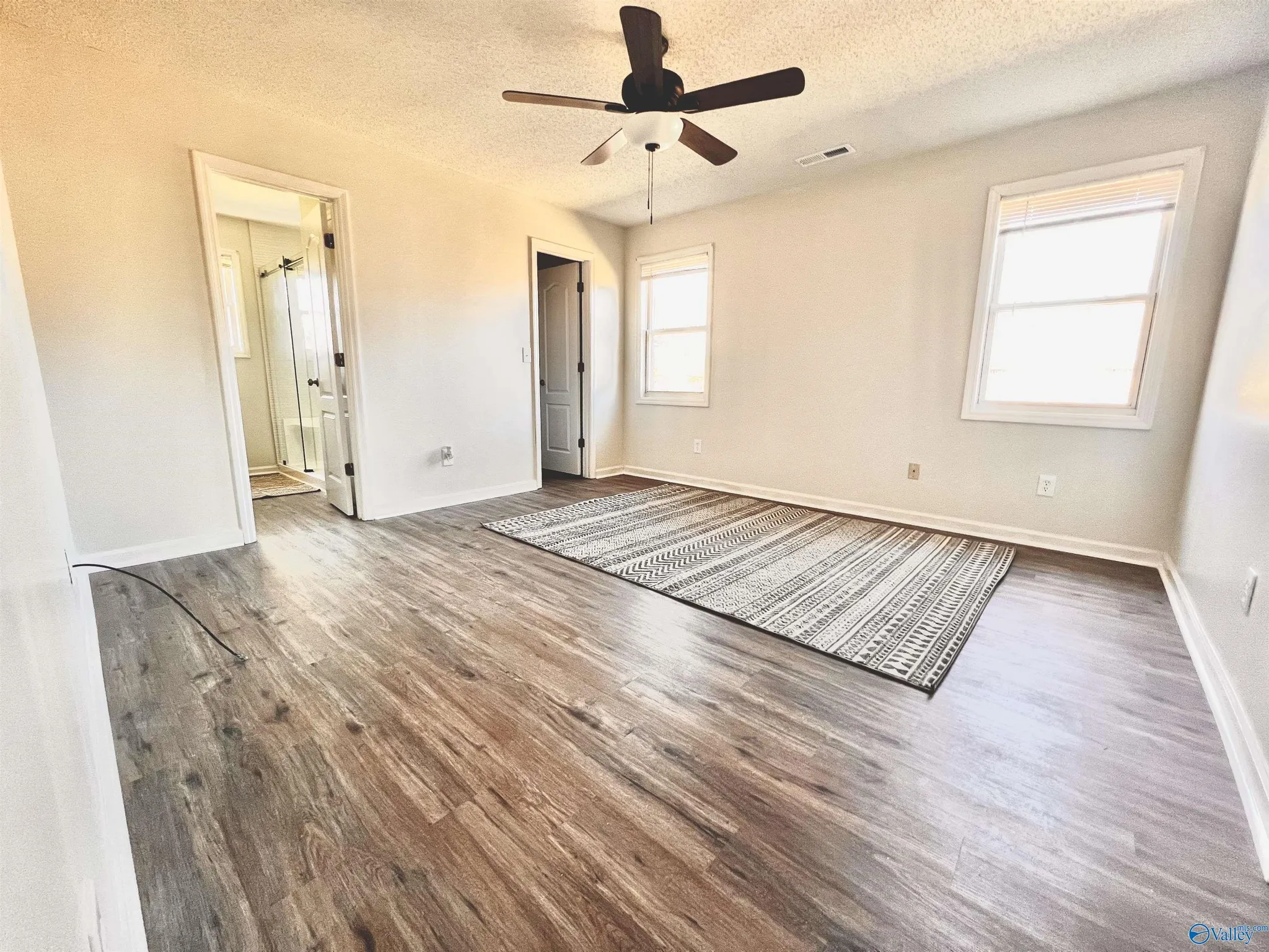 Empty room with wood flooring, white walls, two windows with blinds, a ceiling fan, an open doorway leading to a bathroom, and a patterned rug.