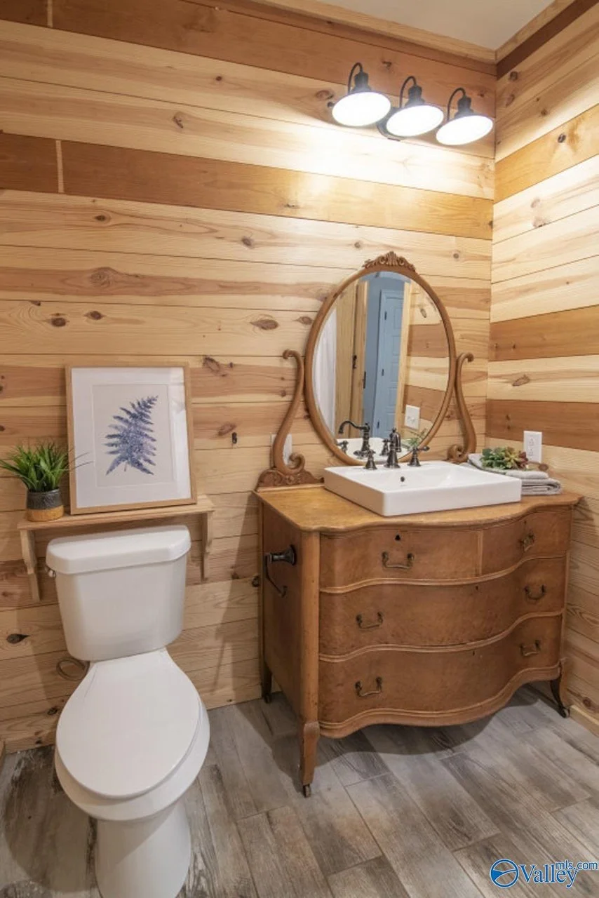 A rustic bathroom with wood-paneled walls, a white toilet, a vintage wooden vanity with a white rectangular sink, a decorative oval mirror, a light fixture with three bulbs, a framed botanical print on a small wooden shelf, and a potted plant.