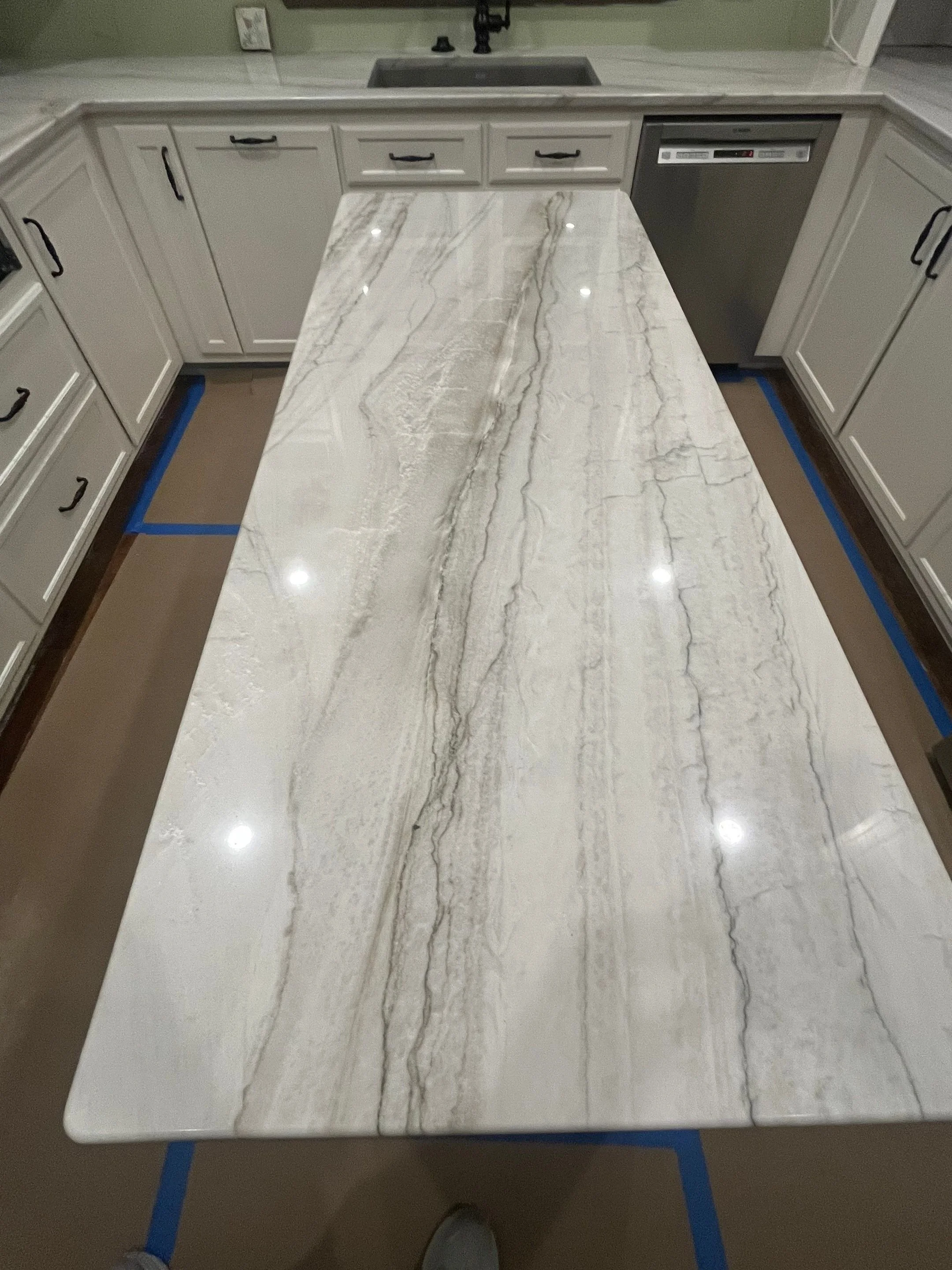 A marble kitchen island with a white and gray veined pattern, surrounded by white cabinets and a dishwasher, with a sink and faucet in the background.