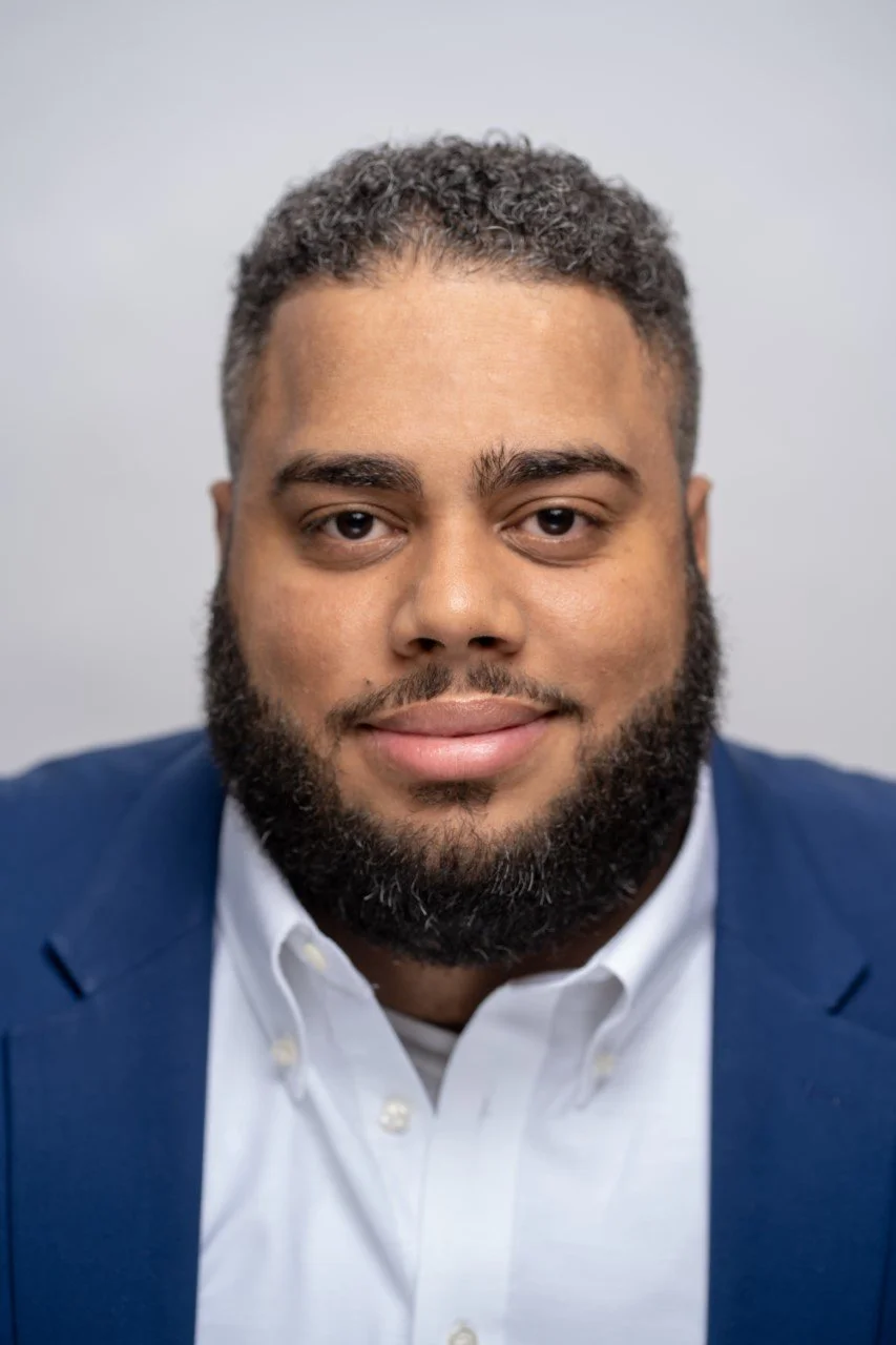 Headshot of a man with a beard and curly hair, wearing a white shirt and blue blazer, against a light gray background.