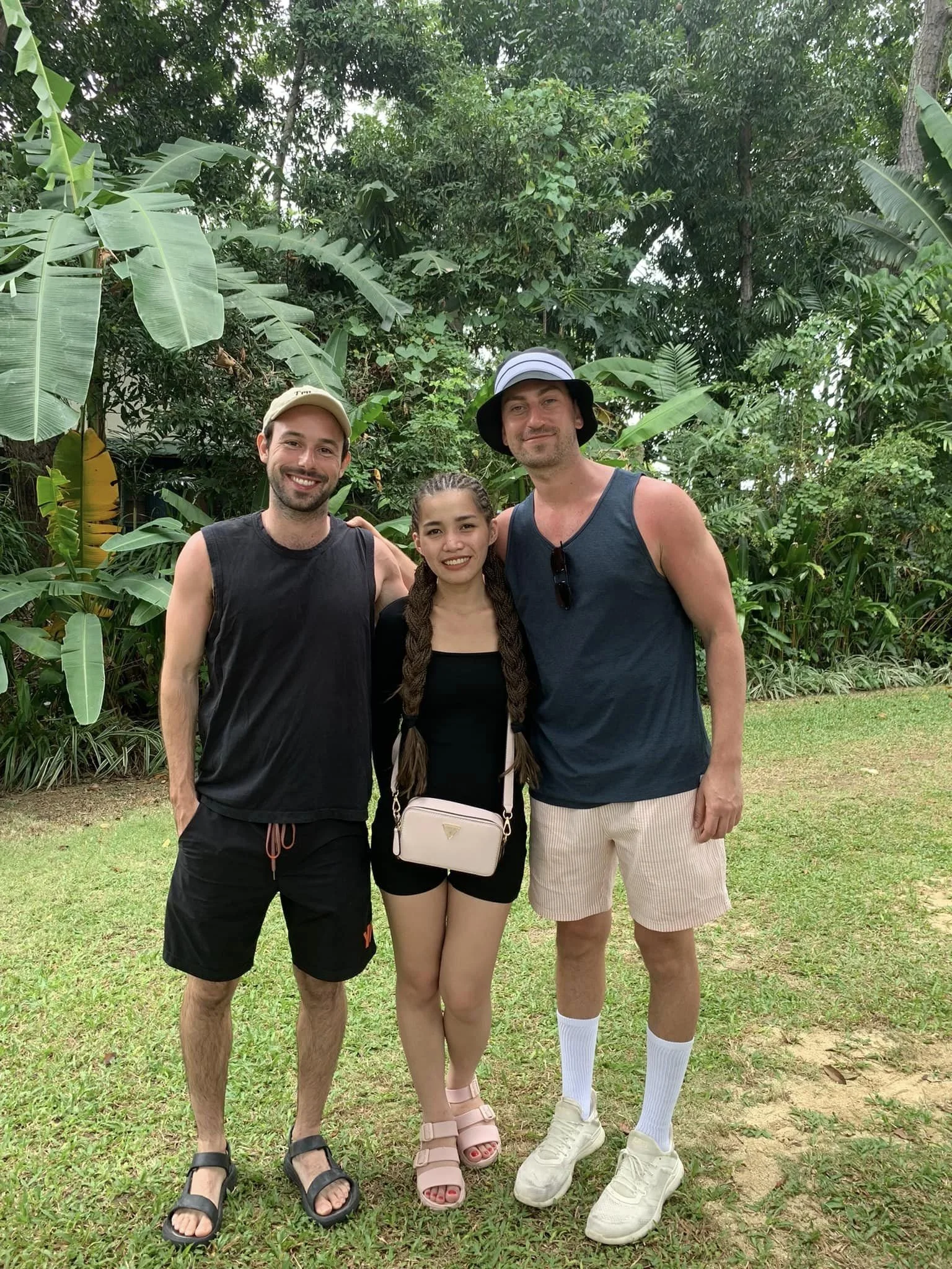 Three people standing together outdoors in a lush green tropical environment, smiling at the camera. The person on the left is wearing a beige cap, sleeveless black shirt, black shorts, and black sandals. The person in the middle is a woman with long braided hair, wearing a black top, black shorts, pink sandals, and carrying a small white purse. The person on the right is wearing a blue tank top, striped shorts, white socks, white sneakers, and a black and white wide-brimmed hat.