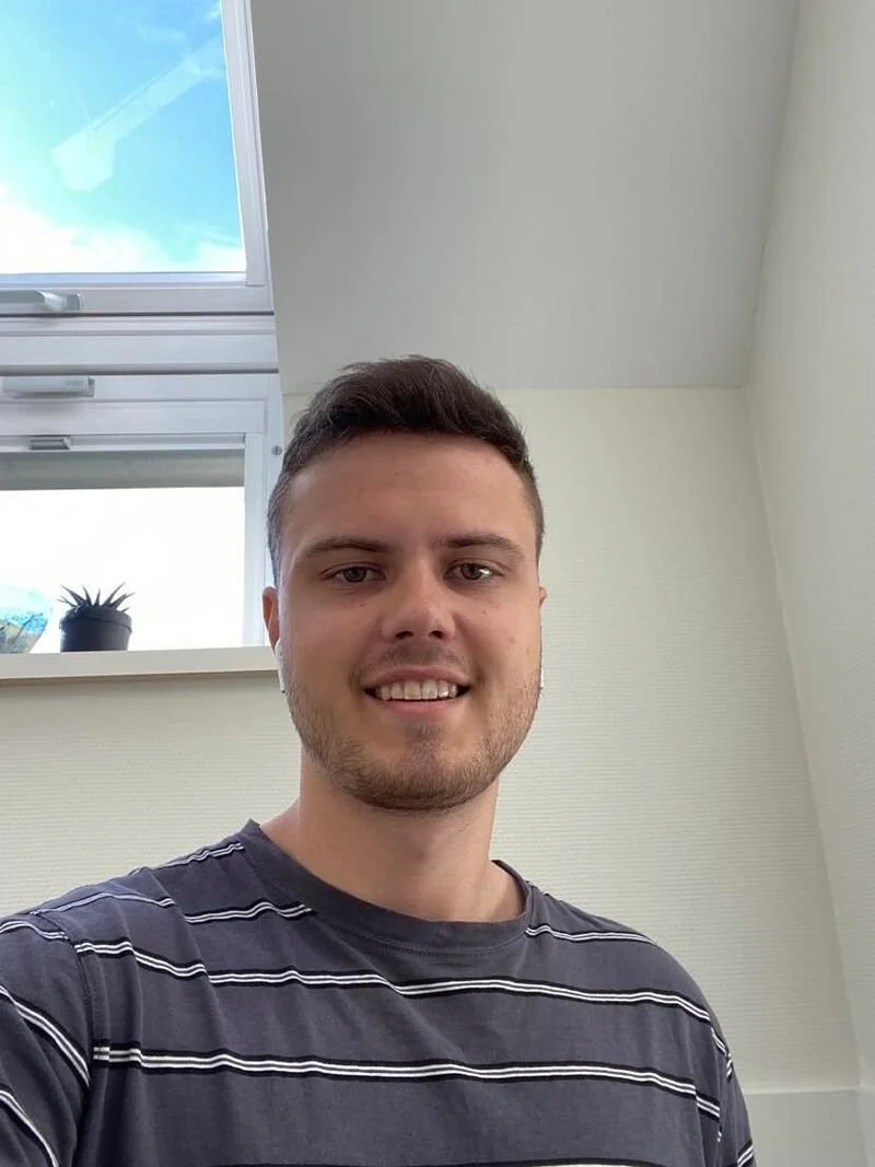 A young man with short dark hair and a light beard smiling, taking a selfie indoors. Behind him is a window with a plant on the sill, and the sky is visible through the window.