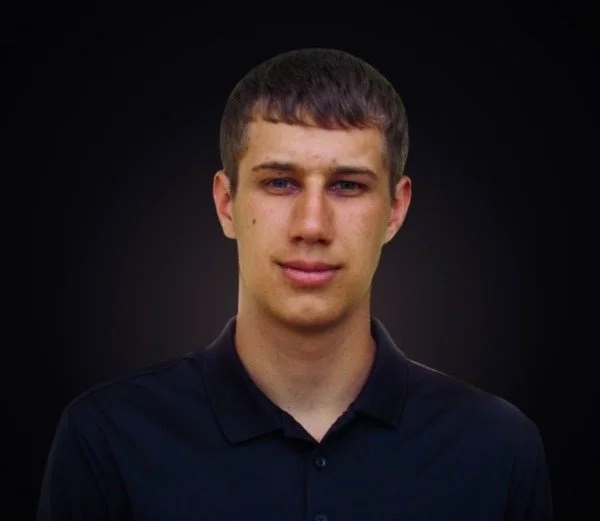 A young man with short brown hair and blue eyes, wearing a black collared shirt, stands against a dark background.
