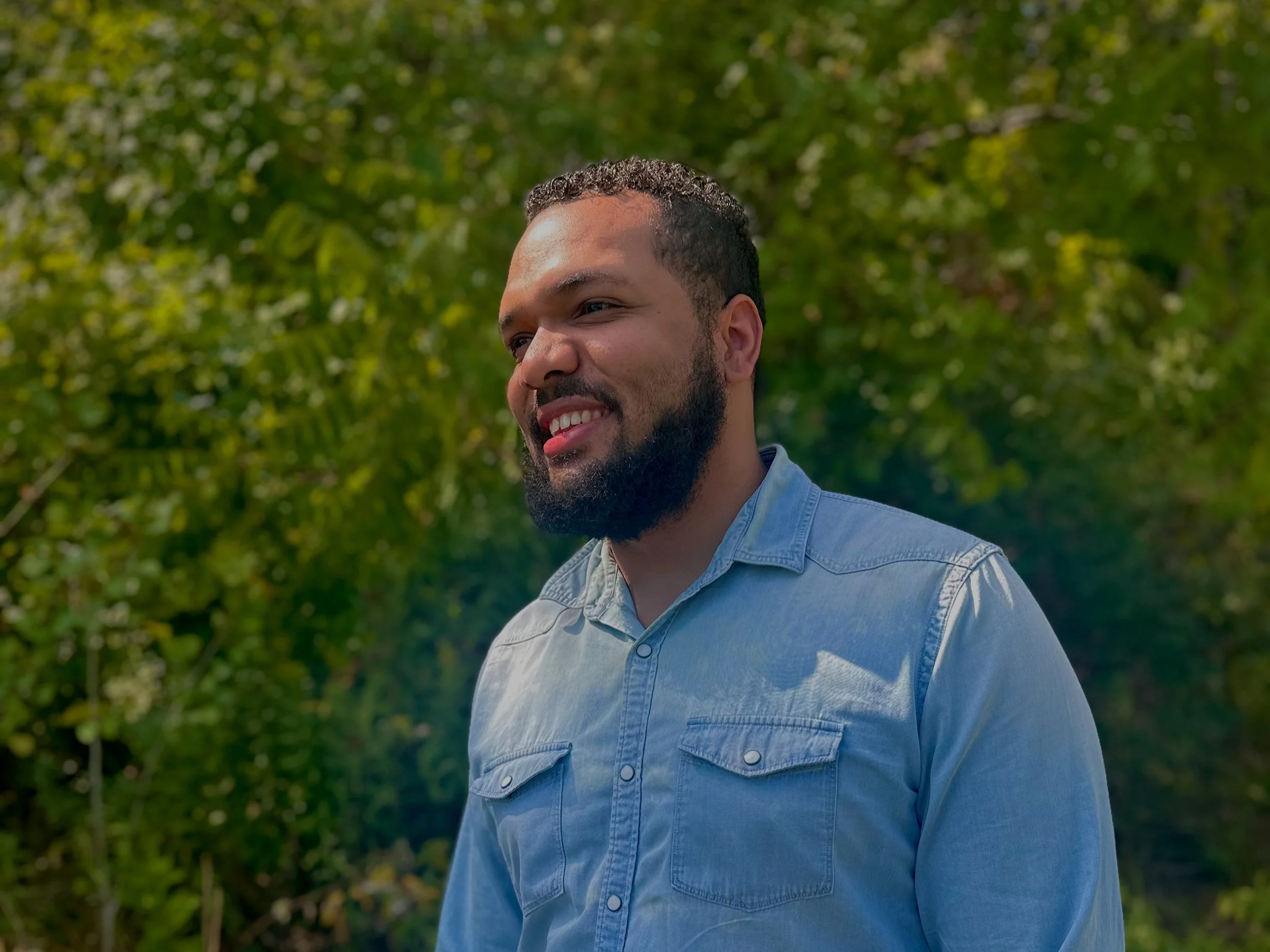 A man with curly hair and a beard, smiling, wearing a light blue denim shirt, standing outdoors with green foliage in the background.