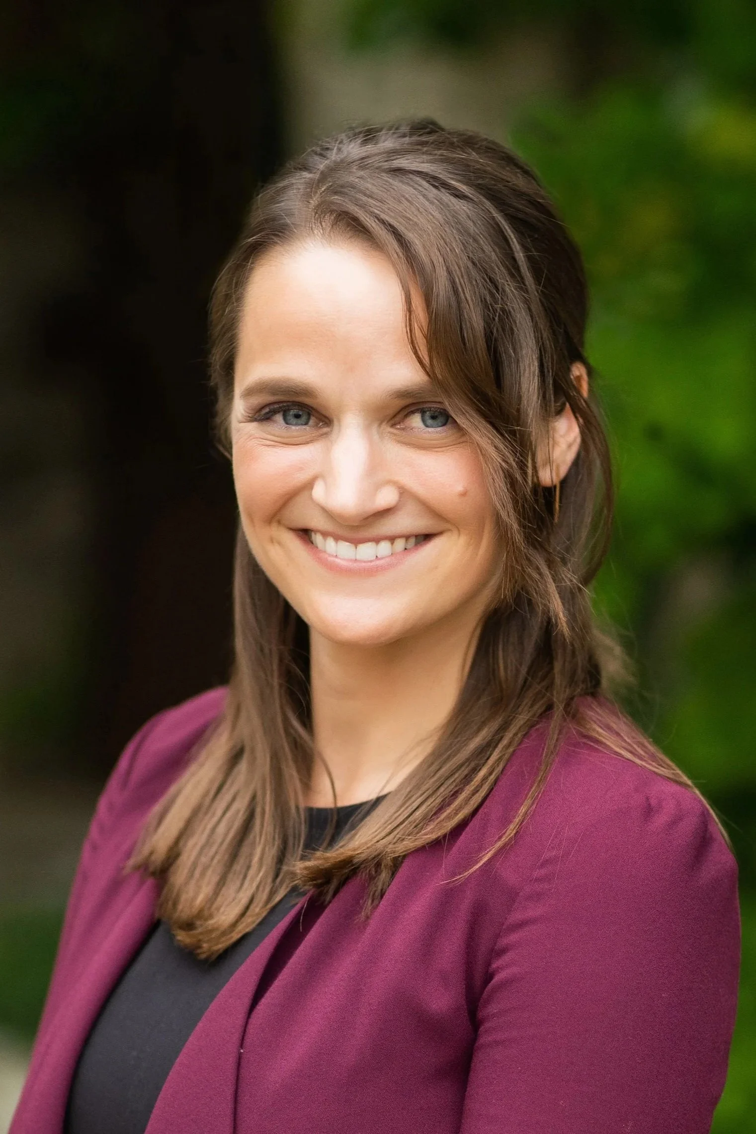 Close-up of a smiling woman with blue eyes and long brown hair, wearing a burgundy blazer over a black top, standing outdoors with greenery in the background.