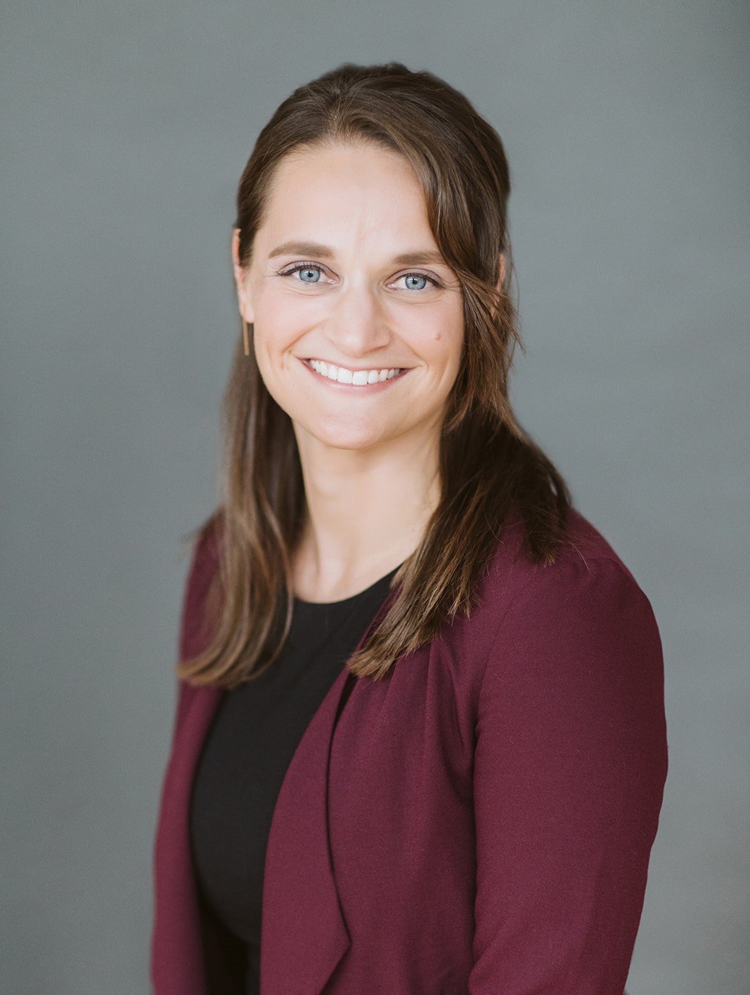 Professional headshot of a woman with shoulder-length brown hair, blue eyes, wearing a black top and a maroon blazer, smiling against a gray background.